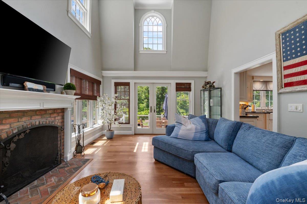 5 Fox Hunt Court Cold Spring Harbor, NY 11724 - Photo 8 of 42 Living area featuring vaulted ceilings, a brick-surround fireplace with a white mantel, wood-finish flooring, multiple windows with wood blinds, and glass French doors