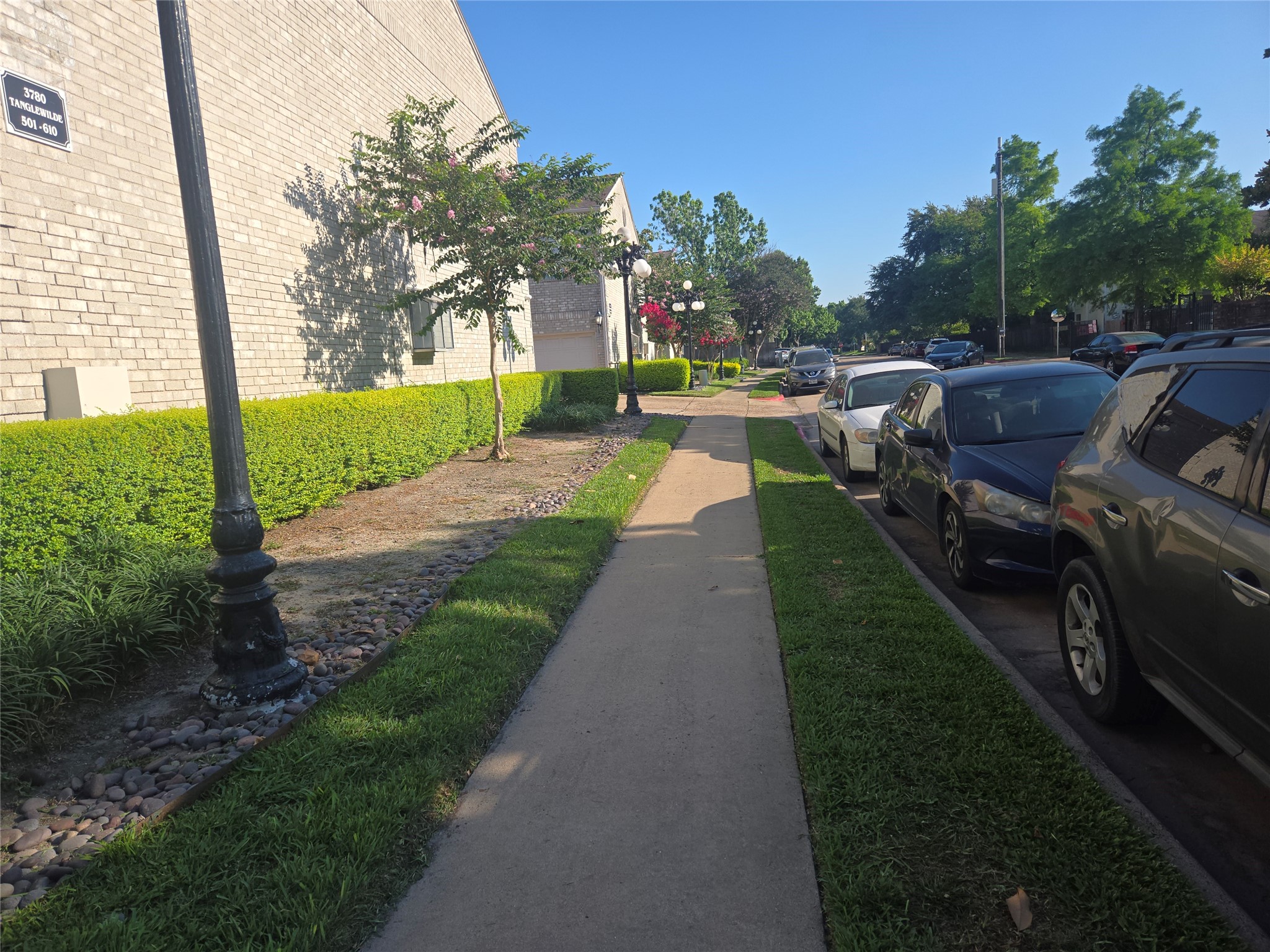 3780 Tanglewilde Street, Unit 501 Houston, TX 77063 - Photo 2 of 26 A well-maintained residential area with a paved sidewalk, neatly trimmed hedges, and parked cars along the street. The area appears peaceful and green, ideal for a pleasant neighborhood walk.