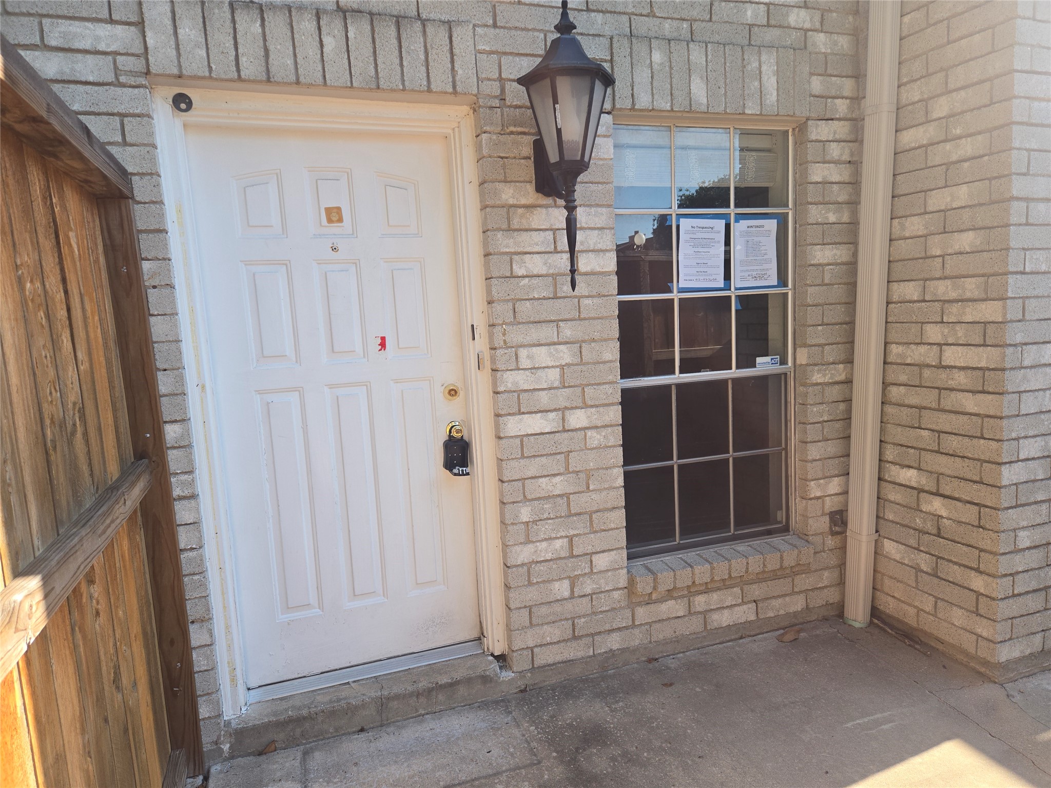 3780 Tanglewilde Street, Unit 501 Houston, TX 77063 - Photo 23 of 26 This photo shows the entrance of a home with a white door and a brick exterior. There is a window to the right with notices posted on it, and a classic outdoor lantern above. A wooden fence is partially visible on the left.