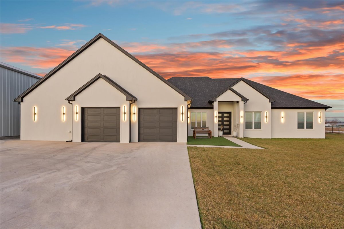 10940 Wedel Cemetery Road Rogers, TX 76569 - Photo 2 of 31 Modern facade with sleek garage doors, a concrete driveway, and ample lawn space set against a vivid sunset.