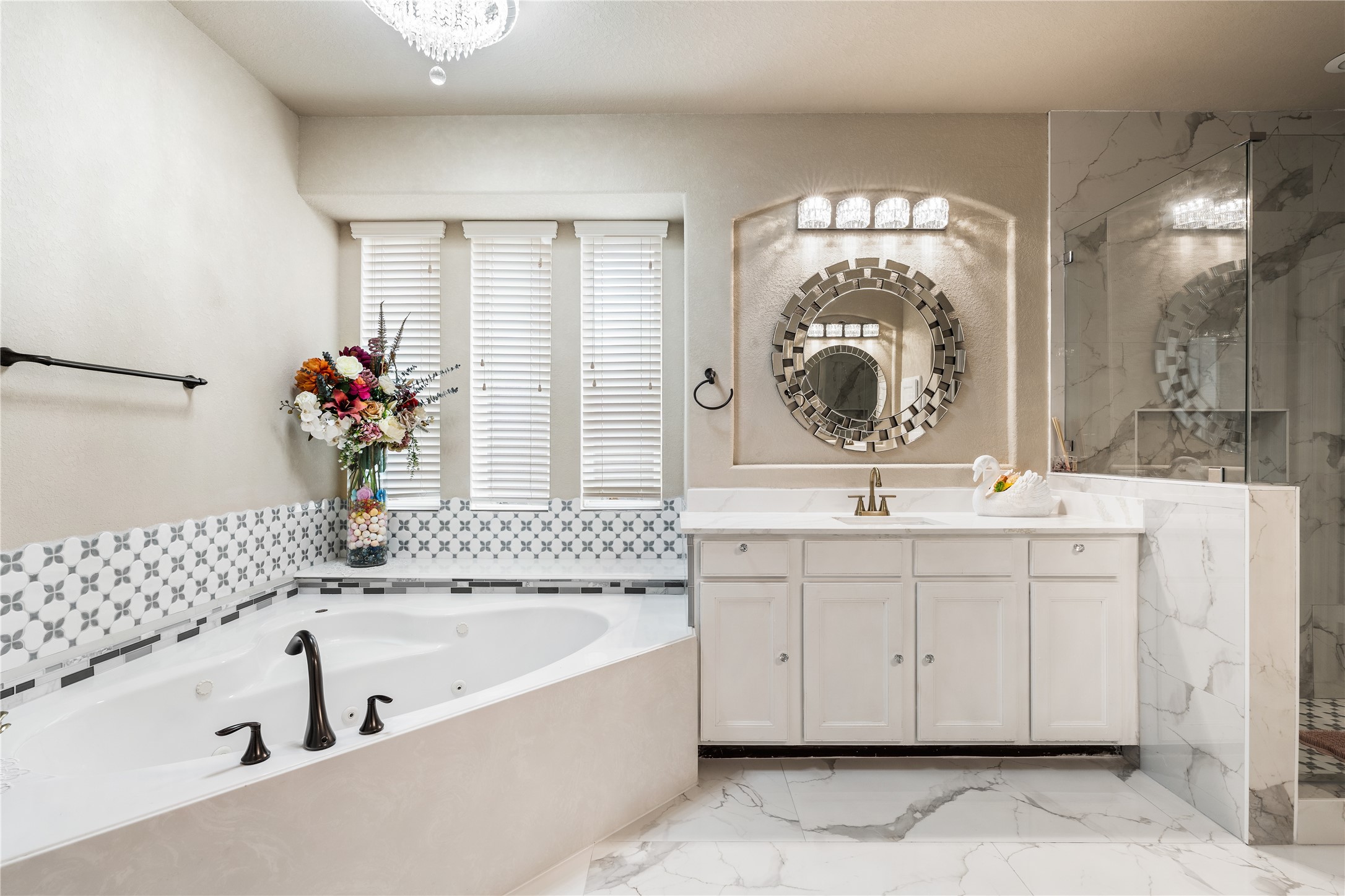 5802 Layton Meadows Lane Spring, TX 77379 - Photo 22 of 42 Full spa view — jetted soaking tub with mosaic tile surround, ornate faucet, crystal chandelier overhead, white vanity with gold hardware, decorative mirror, and the glass shower entry on the right.