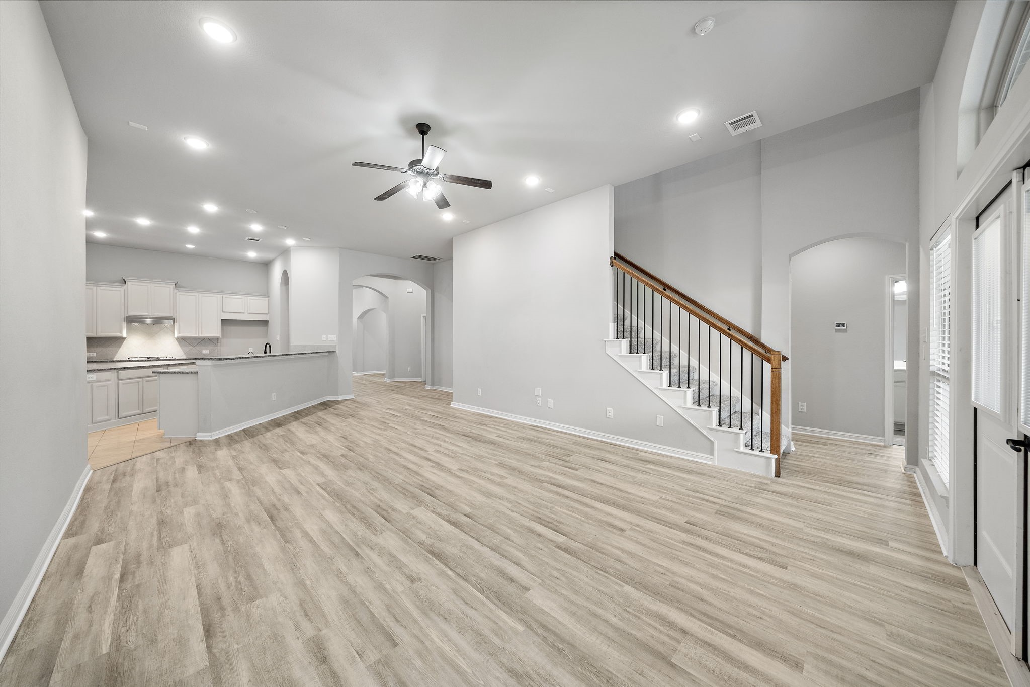 A spacious open-concept living area with light wood flooring, neutral walls, and recessed lighting. A modern kitchen with white cabinetry and a breakfast bar is visible, and a staircase with a sleek railing leads to the upper level. Large windows provide ample natural light.