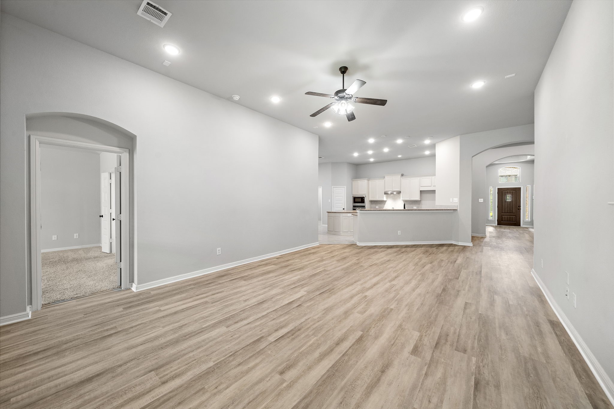 27924 Seger Bnd Trail Spring, TX 77386 - Photo 8 of 21 This spacious open-concept living area features modern wood flooring, a ceiling fan, and ample recessed lighting. It seamlessly connects to a bright kitchen with a large island and white cabinetry. An arched doorway leads to a carpeted room, perfect for a bedroom or office. The entryway is inviting with a view of the front door and windows.