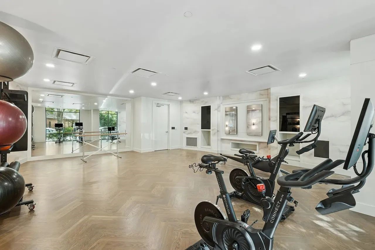2724 Kipling Street, Unit 335 Houston, TX 77098 - Photo 10 of 29 Designed for versatility and mindful movement, the room functions as a multi-purpose fitness hub: Replacing industrial gym tropes with residential elegance, the space features light-toned herringbone wood floors and marble-effect wall paneling that maximize natural light and a sense of openness. A floor-to-ceiling mirrored wall creates visual depth, while integrated, handle-less niches house digital tech and equipment to maintain a minimalist, clutter-free environment.