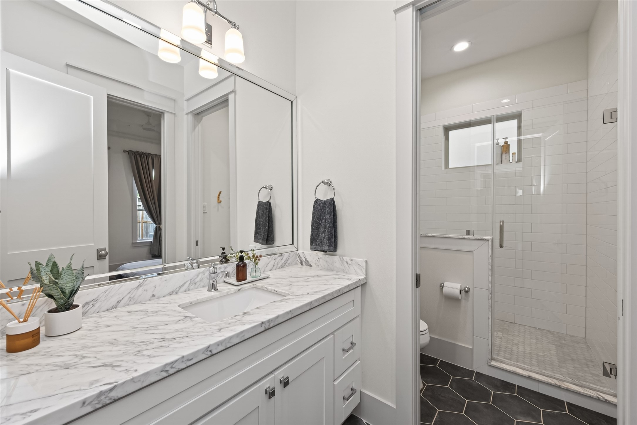 515 West 25th Street Houston, TX 77008 - Photo 13 of 35 En-suite bathroom features ceramic hex tile flooring, Carrara Marble countertop, Kohler toilet, and a beveled mirror above a shaker-style vanity with soft-close drawers, and polished chrome hardware. The frameless glass shower includes a subway tile surround, polished chrome Signature Hardware faucet trim kit, and matching chrome drain, all illuminated by a Park Harbor polished chrome two-bulb vanity light and a frosted clerestory window.