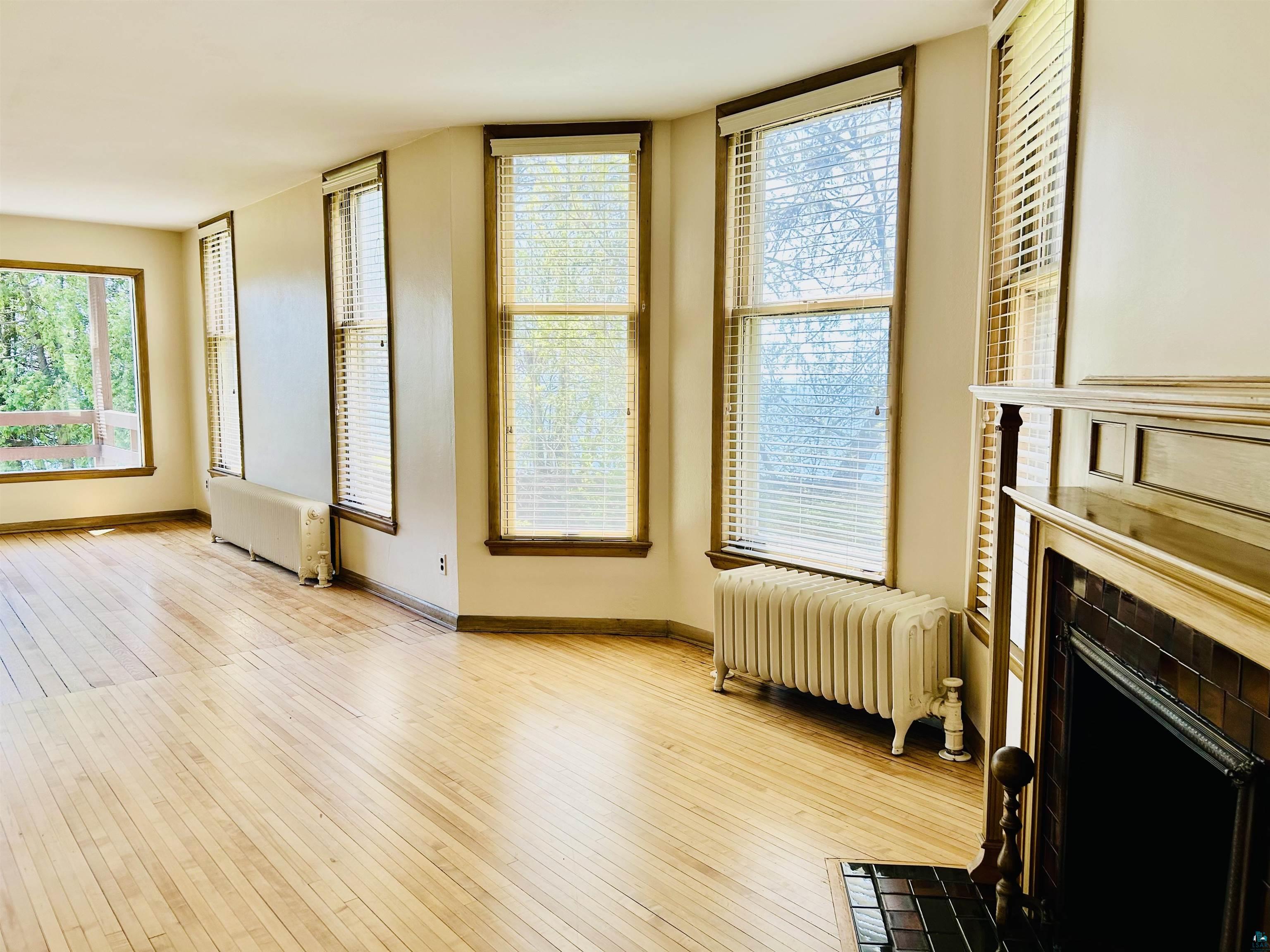 5090 London Road, Unit 1 Duluth, MN 55804 - Photo 18 of 54 Open-concept living area with wood-finish flooring, multiple windows with blinds, cast iron radiators, and a fireplace with a wood mantle and tile surround