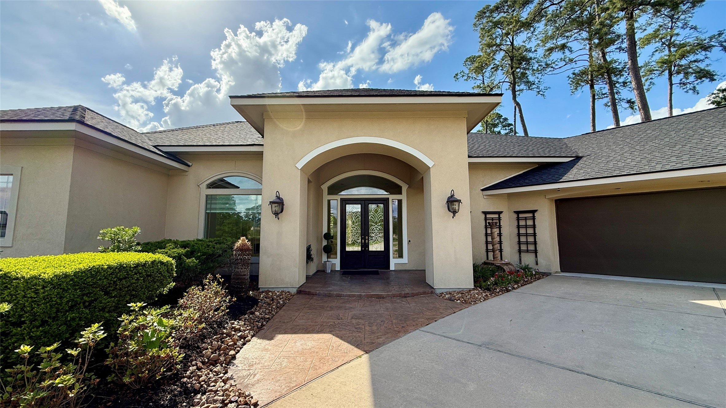 This home features a modern exterior with a welcoming arched entrance, double front doors, and elegant landscaping. It has a spacious driveway leading to a three-car garage, set against a backdrop of tall trees.