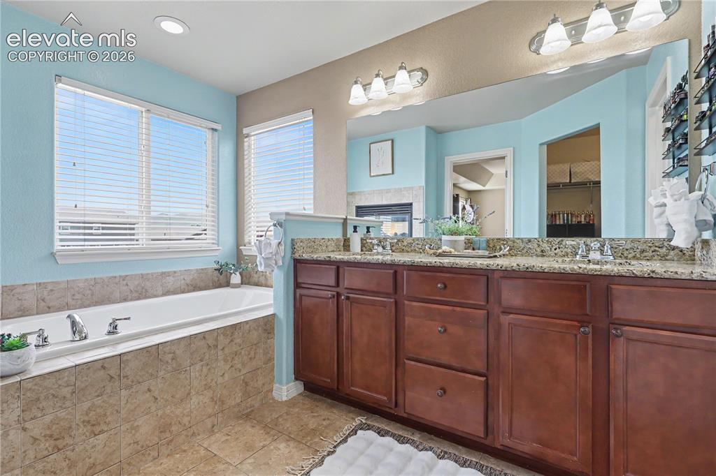 6598 Alliance Loop Colorado Springs, CO 80925 - Photo 17 of 39 Bathroom featuring a built-in soaking tub with tiled surround, dual windows, a double vanity with granite countertop, and wood-finish cabinetry