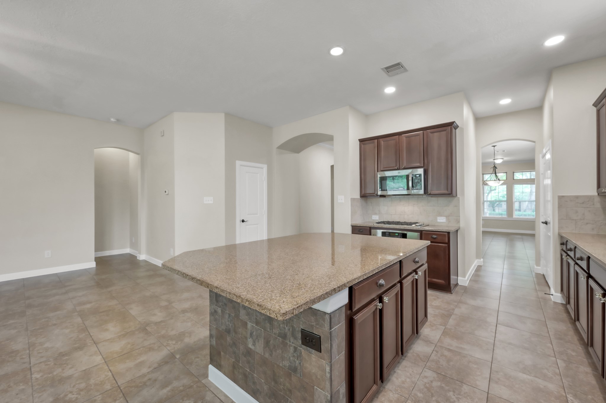 31507 Boulder Cliff Lane Spring, TX 77386 - Photo 19 of 45 This photo showcases a spacious, modern kitchen with dark wood cabinetry, a large central island with a stone countertop, and stainless steel appliances. The open-concept design extends into a bright dining area with tile flooring and recessed lighting, offering a perfect blend of style and functionality.