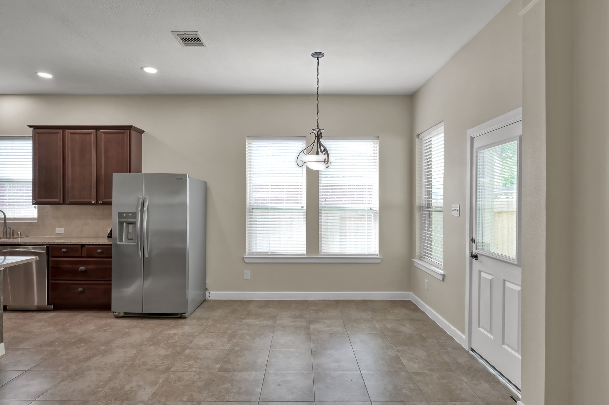 31507 Boulder Cliff Lane Spring, TX 77386 - Photo 21 of 45 This photo showcases a bright kitchen-dining area with large windows, neutral walls, and tiled flooring. It features dark wood cabinets, a stainless steel refrigerator, and a hanging light fixture. There is a door leading outside, enhancing natural light.