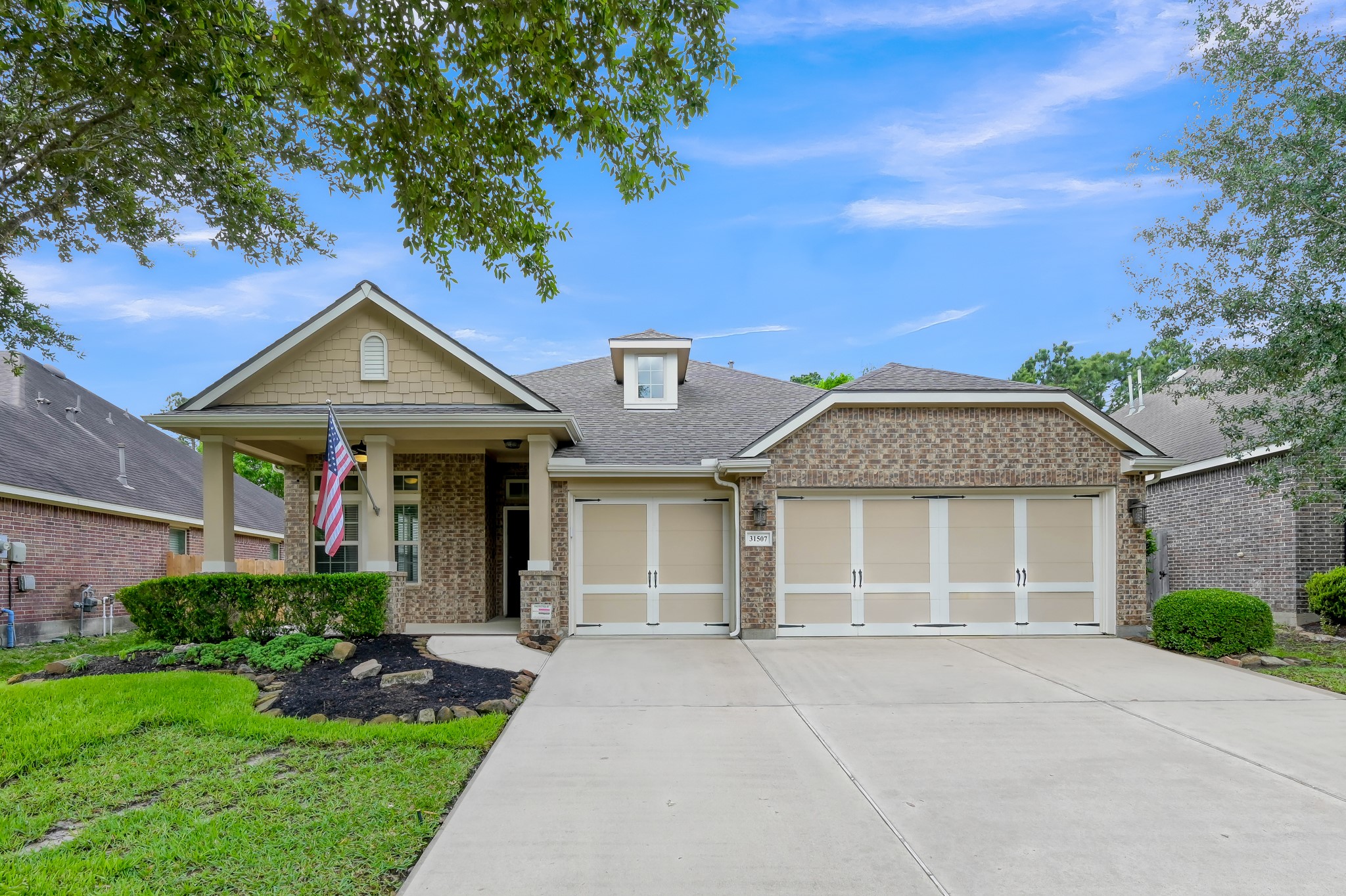 31507 Boulder Cliff Lane Spring, TX 77386 - Photo 45 of 45 This photo showcases a charming, single-story home with a brick facade and a three-car garage. The well-maintained front yard features manicured bushes and a tree, enhancing the home's curb appeal. A welcoming front porch with a flag adds to its inviting appearance.