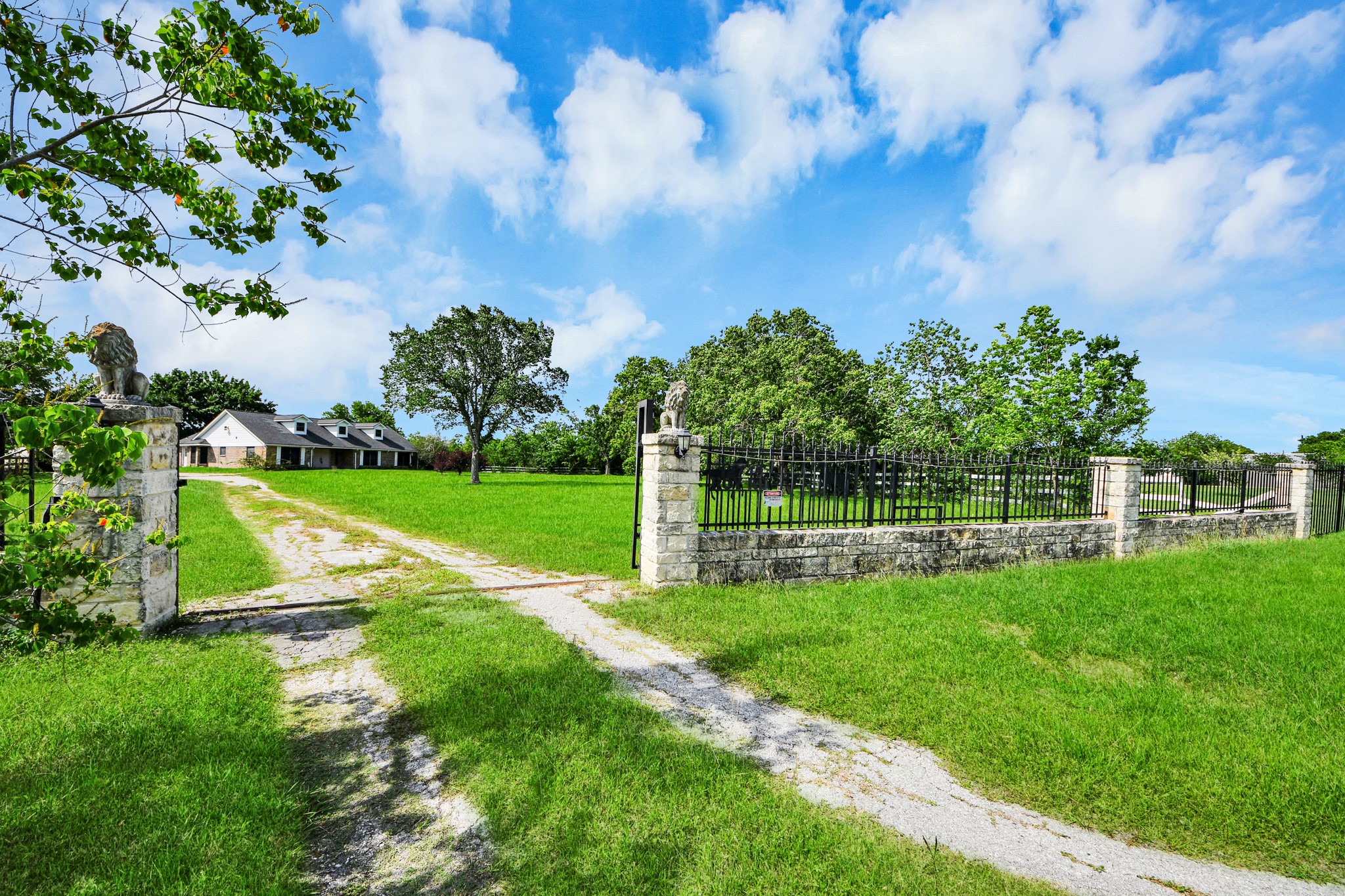 12746 Roy Road Pearland, TX 77581 - Photo 1 of 47 This photo features a charming country home set on a spacious green lawn. A long driveway leads to the house, passing through a stone gate with decorative statues. The property is surrounded by mature trees and open sky, offering a sense of privacy and tranquility.