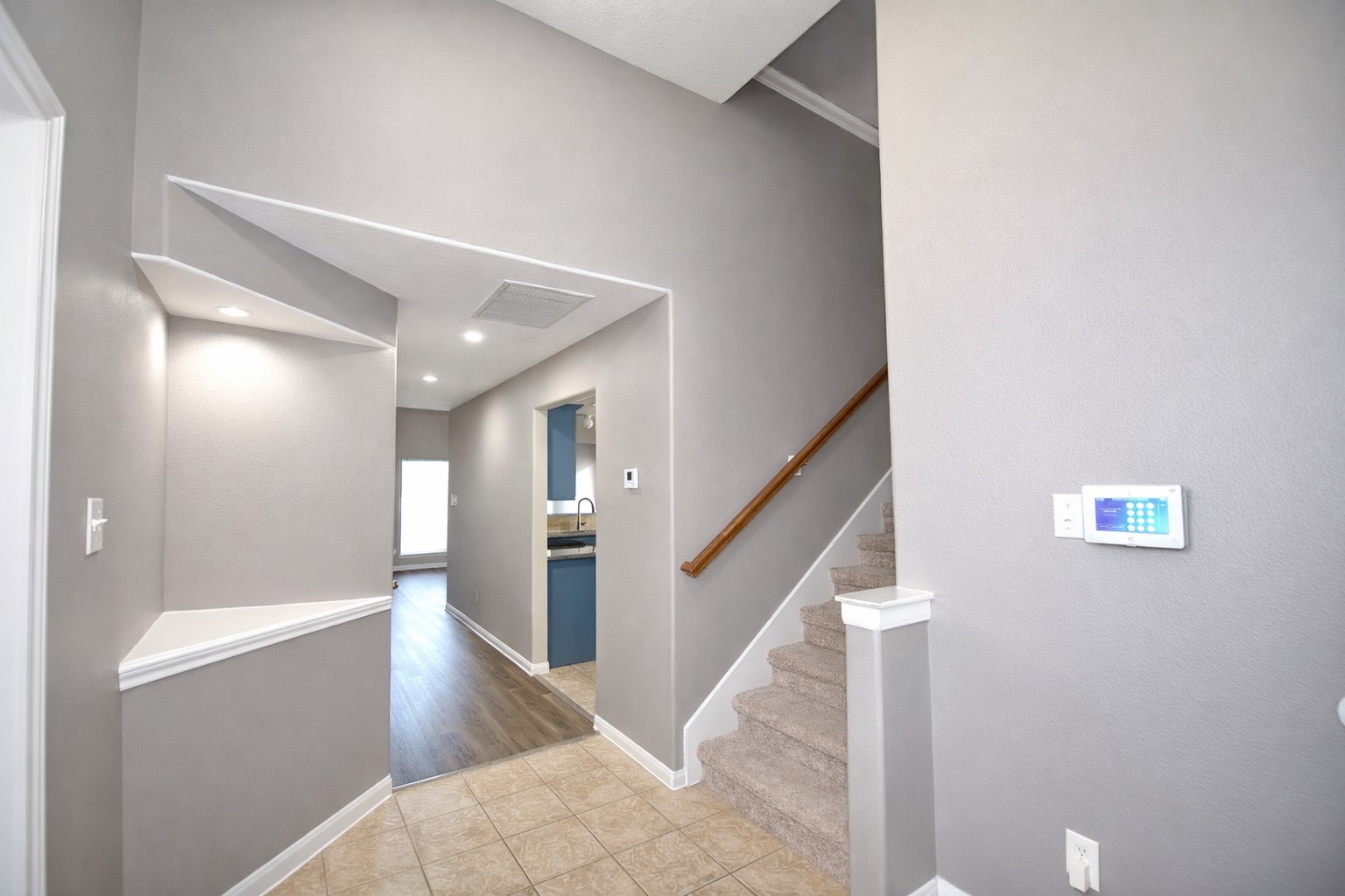 9611 Babbling Spring Court Tomball, TX 77375 - Photo 4 of 31 This photo showcases a modern home entryway with neutral gray walls, tile flooring, and a staircase leading to the upper level. Recessed lighting illuminates the hall, which leads to a kitchen with visible wood and tile flooring. The space includes a wall-mounted control panel.