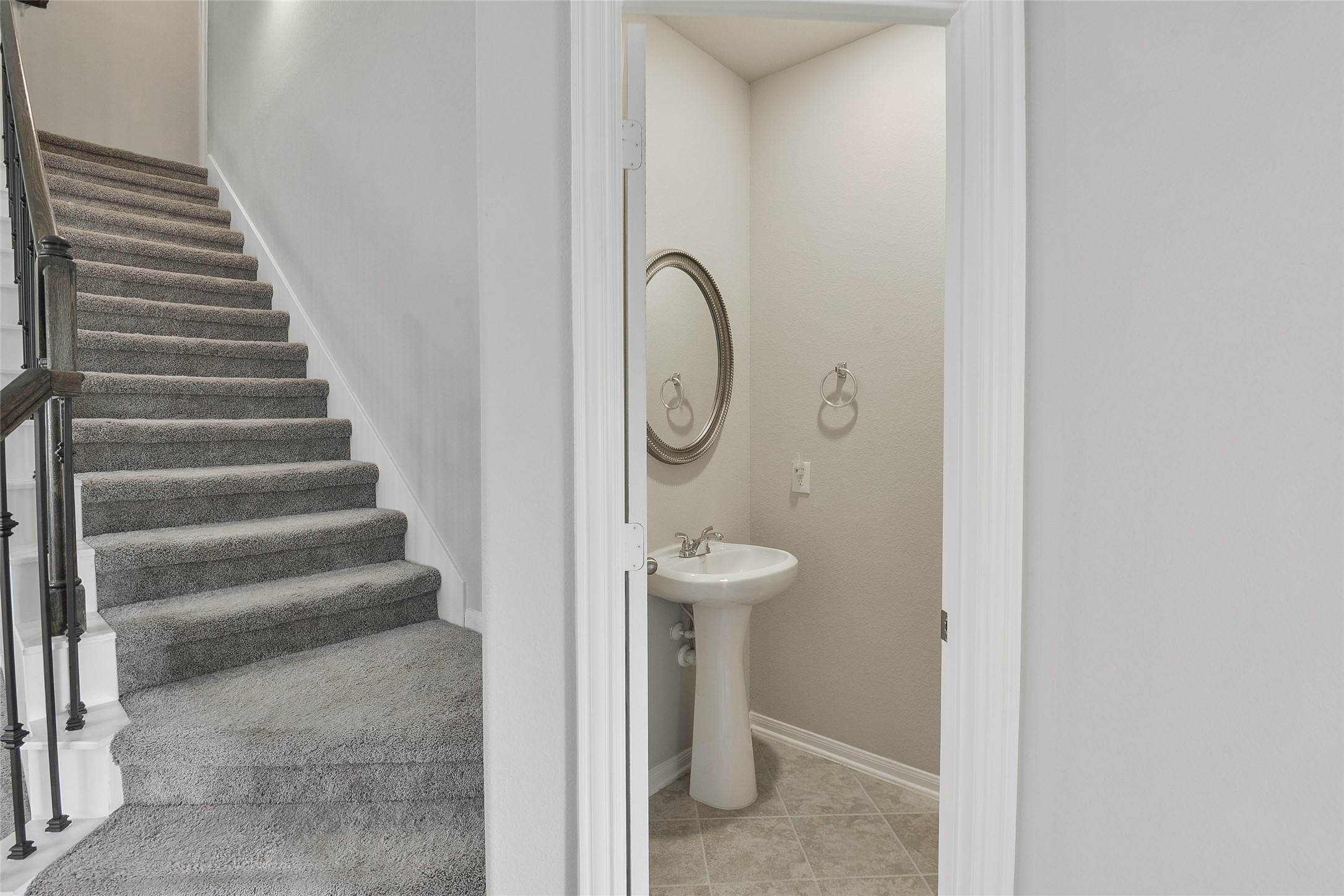 614 Pequin Road Crosby, TX 77532 - Photo 22 of 43 This features a carpeted staircase with dark railing leading upstairs. Adjacent is a small, modern half-bathroom with a pedestal sink and circular mirror, set on tiled flooring.