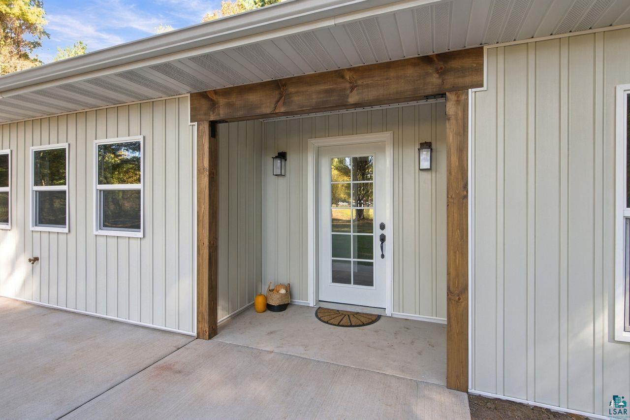65352 Adler Road Marengo, WI 54855 - Photo 6 of 24 Board and batten siding with white trim, a covered entryway with exposed wood beams, a glass-paneled front door, and exterior sconce lighting