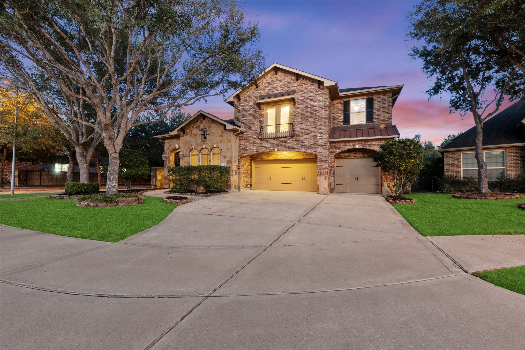 5827 Amherst Farms Lane Fulshear, TX 77441 - Photo 37 of 48 Twilight elegance highlights this stunning two-story home, where warm lighting showcases the brick and stone façade. A spacious driveway, three-car garage, mature trees, and a charming Juliet balcony create unforgettable curb appeal against a Texas sunset sky.