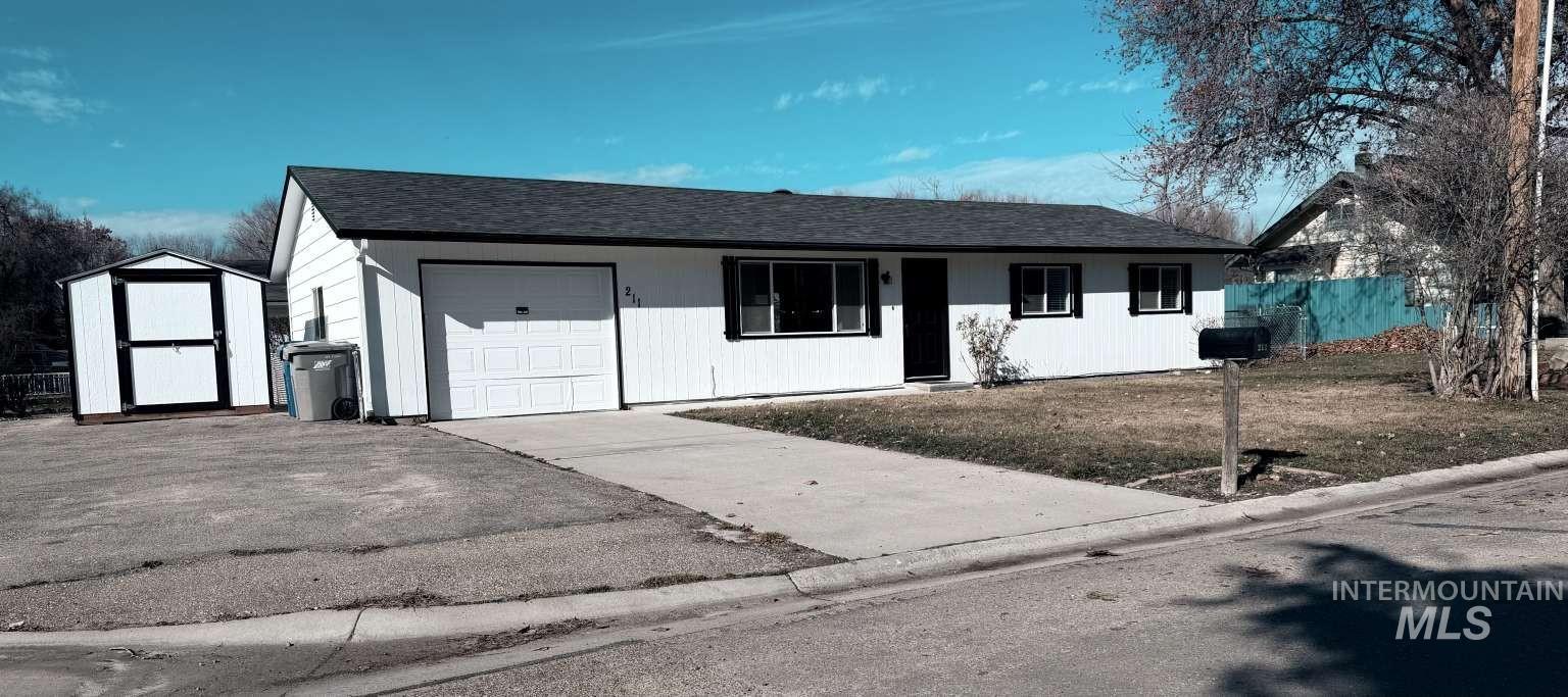 Ranch-style home with a shingled roof, concrete driveway, a garage, and a storage shed