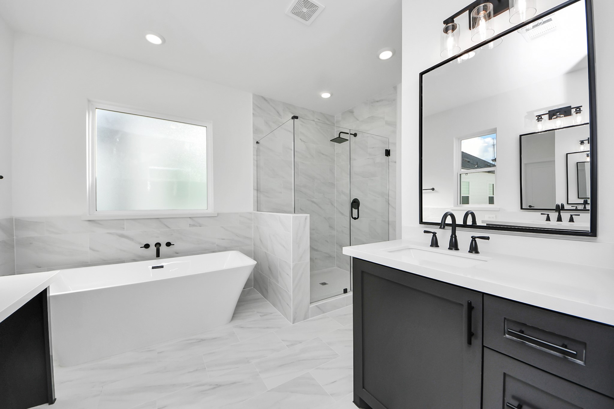 723 Marcella Street, Unit D Houston, TX 77091 - Photo 15 of 22 This modern bathroom features a sleek design with a free standing bathtub, a glass-enclosed shower, and stylish fixtures. The space is brightened by natural light from large windows and has elegant marble-like tiles for a luxurious feel.
