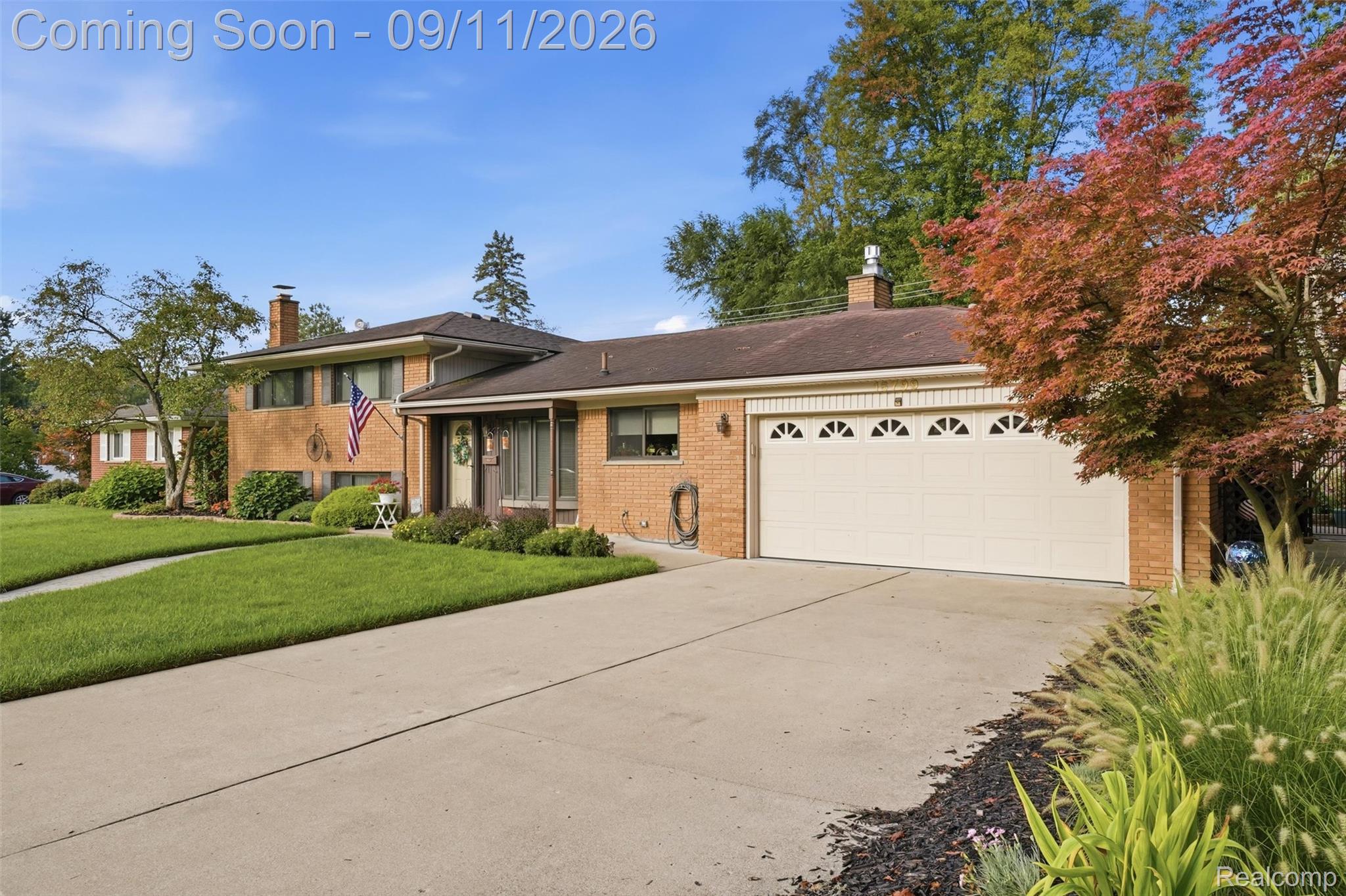 16799 Farmington Road Livonia, MI 48154 - Photo 4 of 71 Brick exterior featuring a two-car garage with arched window accents, a concrete driveway, and established landscaping with mature trees