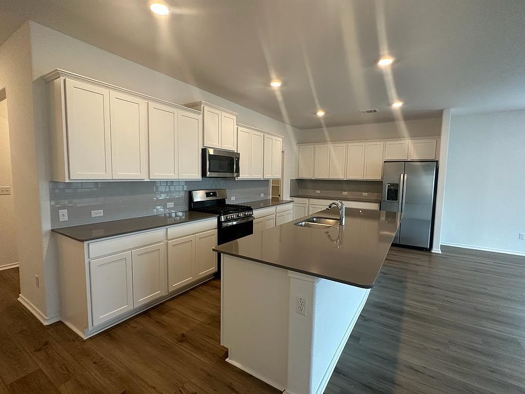 160 River Rise Road Kyle, TX 78640 - Photo 29 of 35 Open-concept kitchen featuring white shaker-style cabinetry, dark countertops, a large island with an undermount sink, stainless steel appliances, and wood-finish flooring