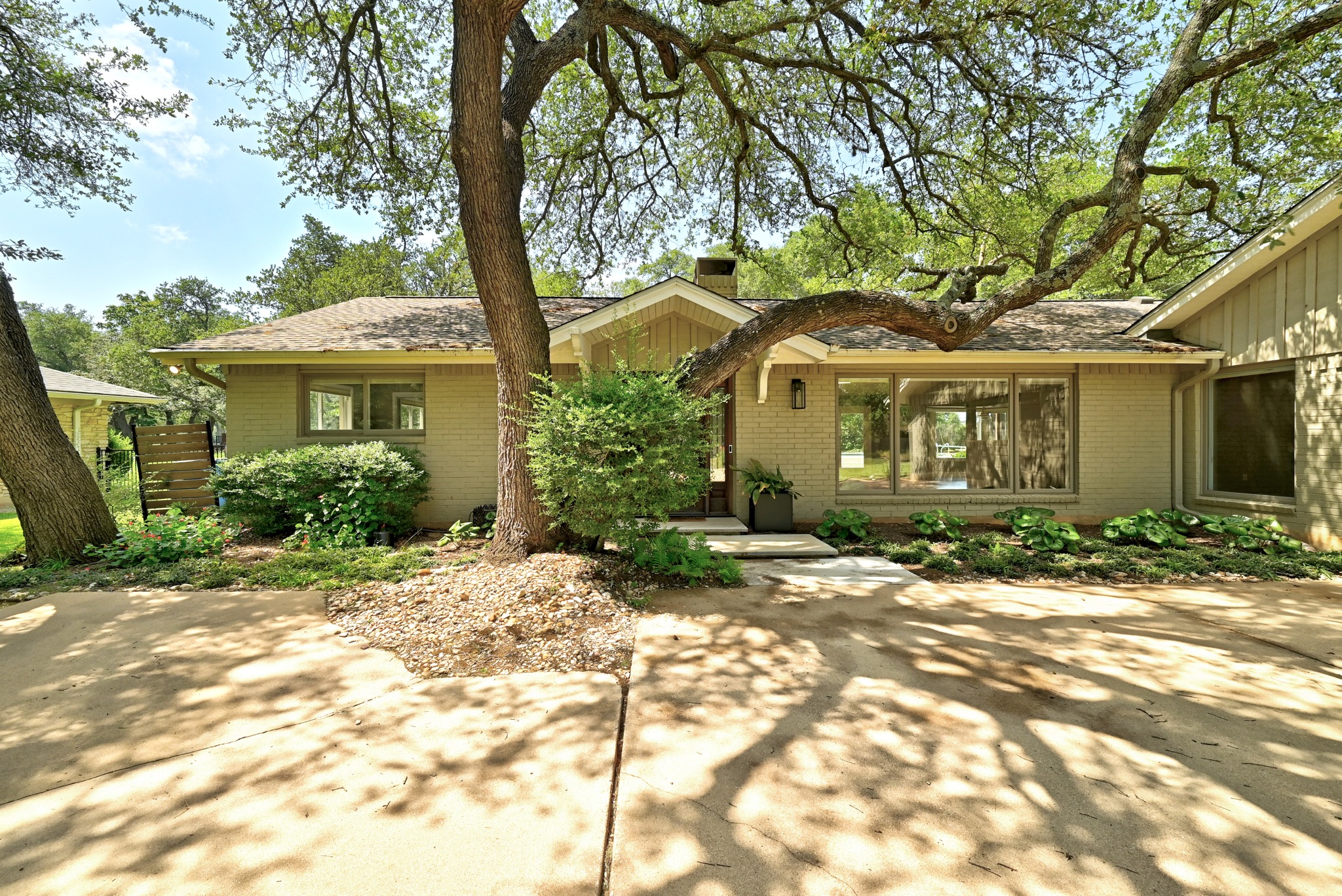 Mature trees canopy the entry to this inviting residence, featuring brick siding and ample front-facing windows.