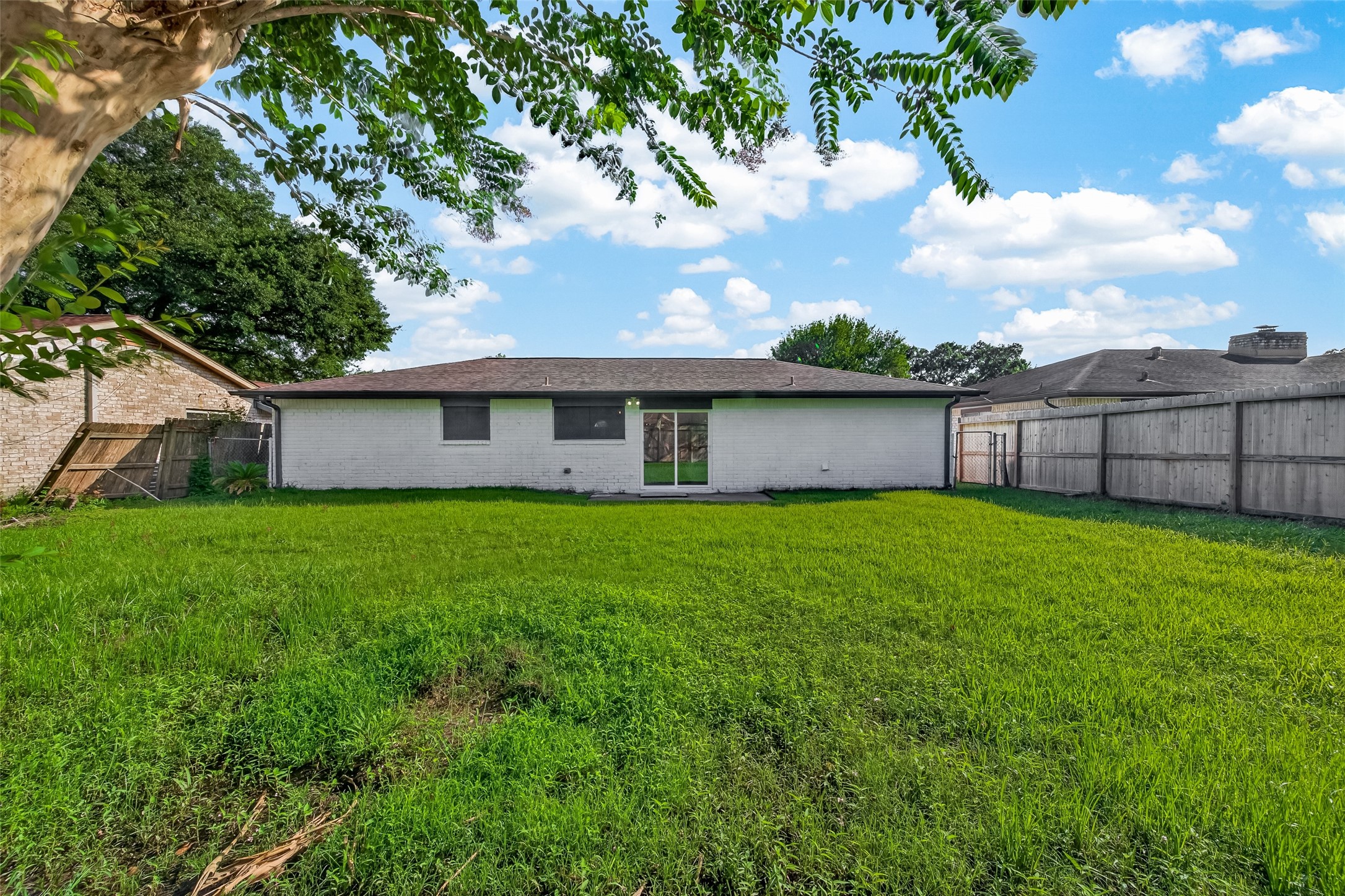 16619 Tibet Road Friendswood, TX 77546 - Photo 27 of 29 This photo shows a spacious backyard with a well-maintained lawn, bordered by a wooden fence. The back of the house has a simple, white exterior with a central sliding glass door, providing easy access to the yard. A tree in the foreground adds a touch of greenery.
