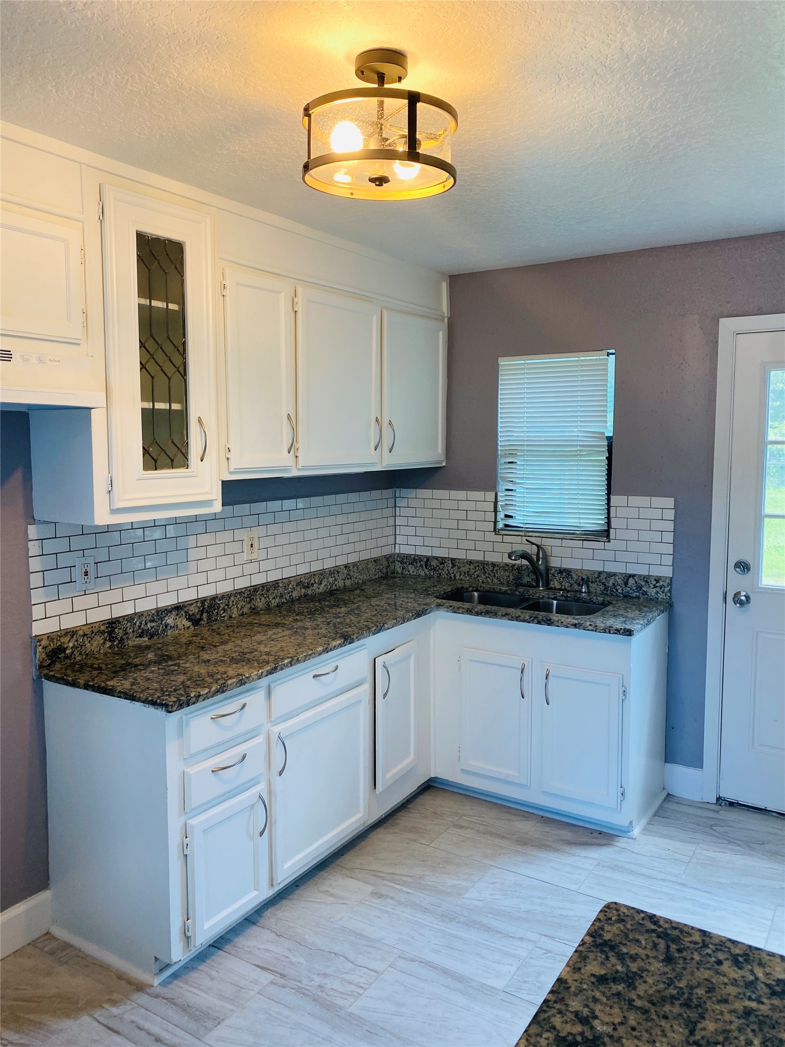 7547 Laura Koppe Road Houston, TX 77028 - Photo 5 of 11 Bright kitchen with white cabinetry, granite countertops, and a modern light fixture. Features a subway tile backsplash and tile flooring, with a window and door providing natural light.