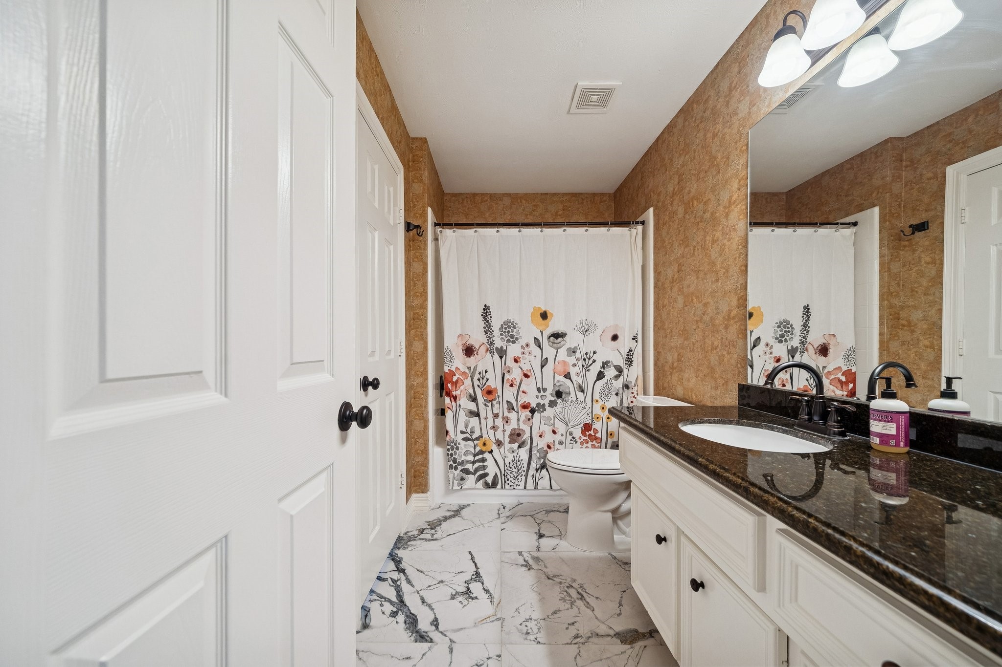 2114 Lytham Lane Katy, TX 77450 - Photo 20 of 29 Guest Bath #2. This bathroom features a modern, elegant design with a dark granite countertop, white cabinetry, and a large mirror. The shower curtain adds a pop of color with floral patterns, and the space is complemented by stylish lighting and marble-look flooring.