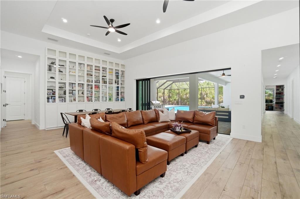 522 31st Street Southwest Naples, FL 34117 - Photo 7 of 38 Spacious living area featuring light wood-finish flooring, a tray ceiling with recessed lighting and ceiling fans, and an expansive built-in white cabinetry wall with glass-fronted upper sections