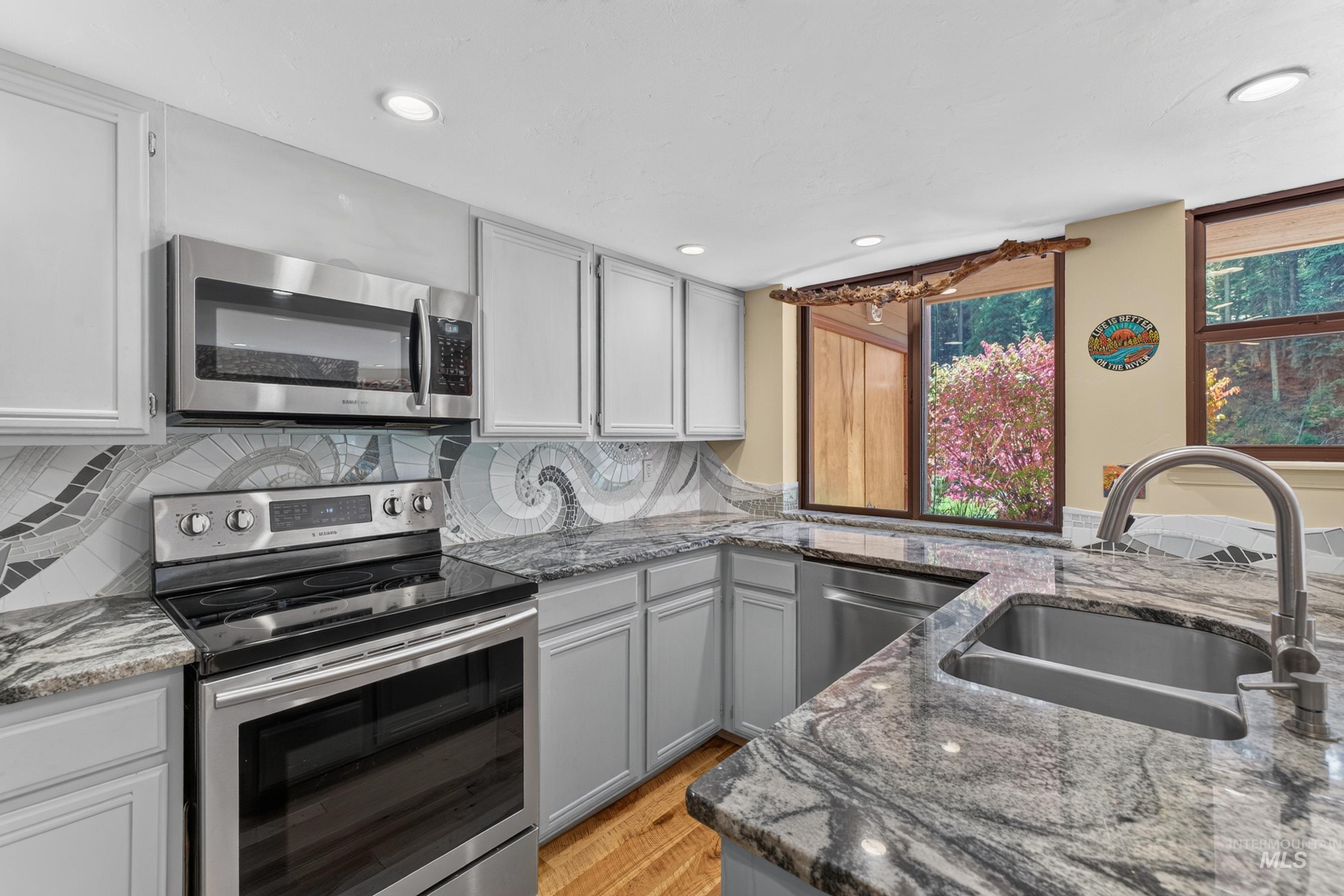 Kitchen featuring gray shaker-style cabinetry, granite countertops, patterned tile backsplash, stainless steel appliances, and a double-basin sink