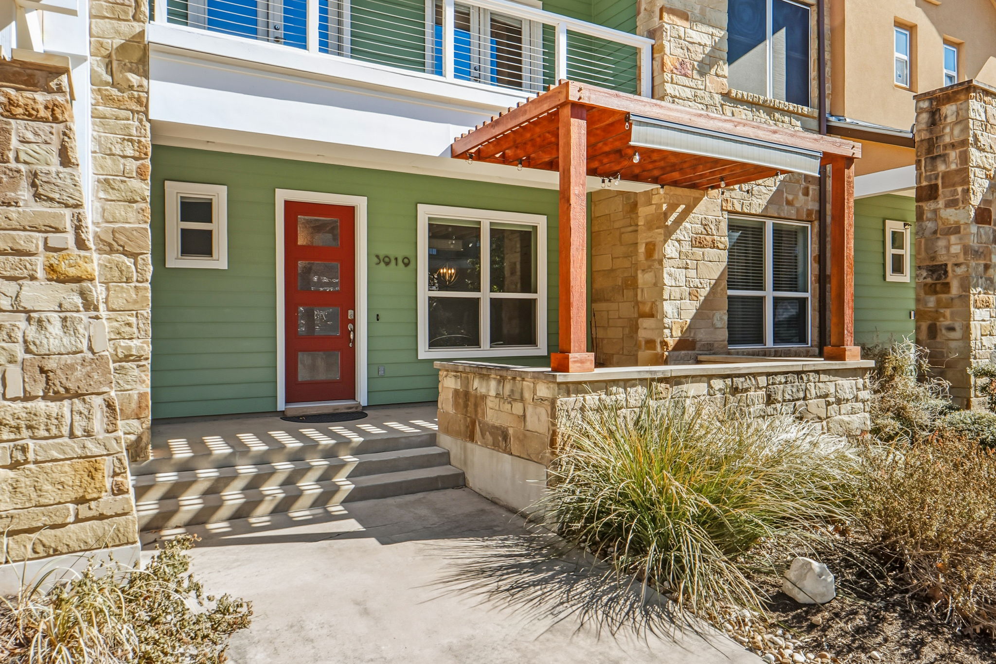 3919 Vaughan Street Austin, TX 78723 - Photo 4 of 40 A vibrant red entry door beckons, framed by verdant siding and natural stone accents, with a welcoming pergola.