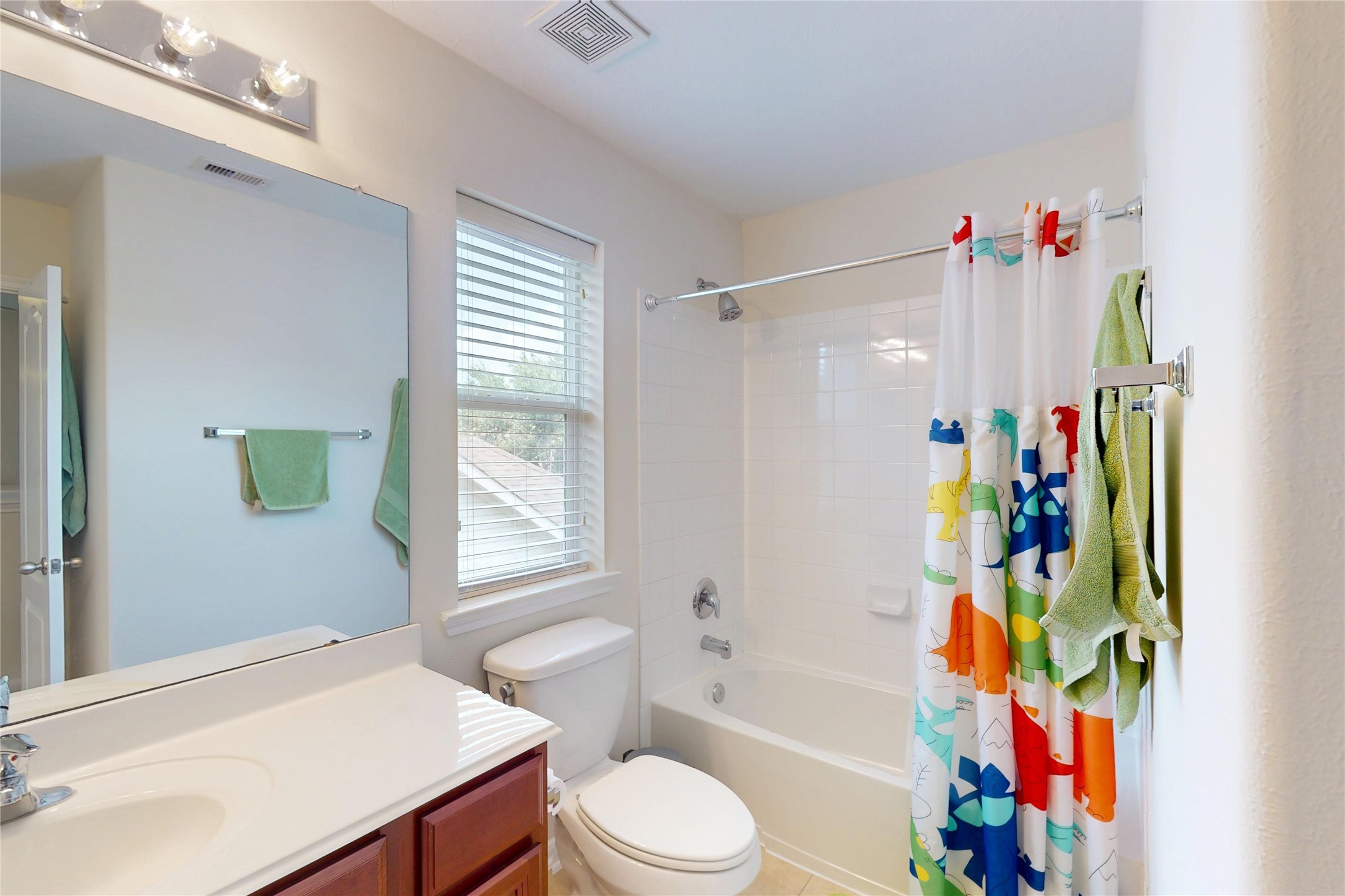 17402 Rainer Valley Lane Humble, TX 77346 - Photo 21 of 33 Bright and inviting bathroom with a modern vanity, large mirror, and a window for natural light. Features a bathtub with a colorful shower curtain, white tiles, and light walls, creating a fresh and clean atmosphere.