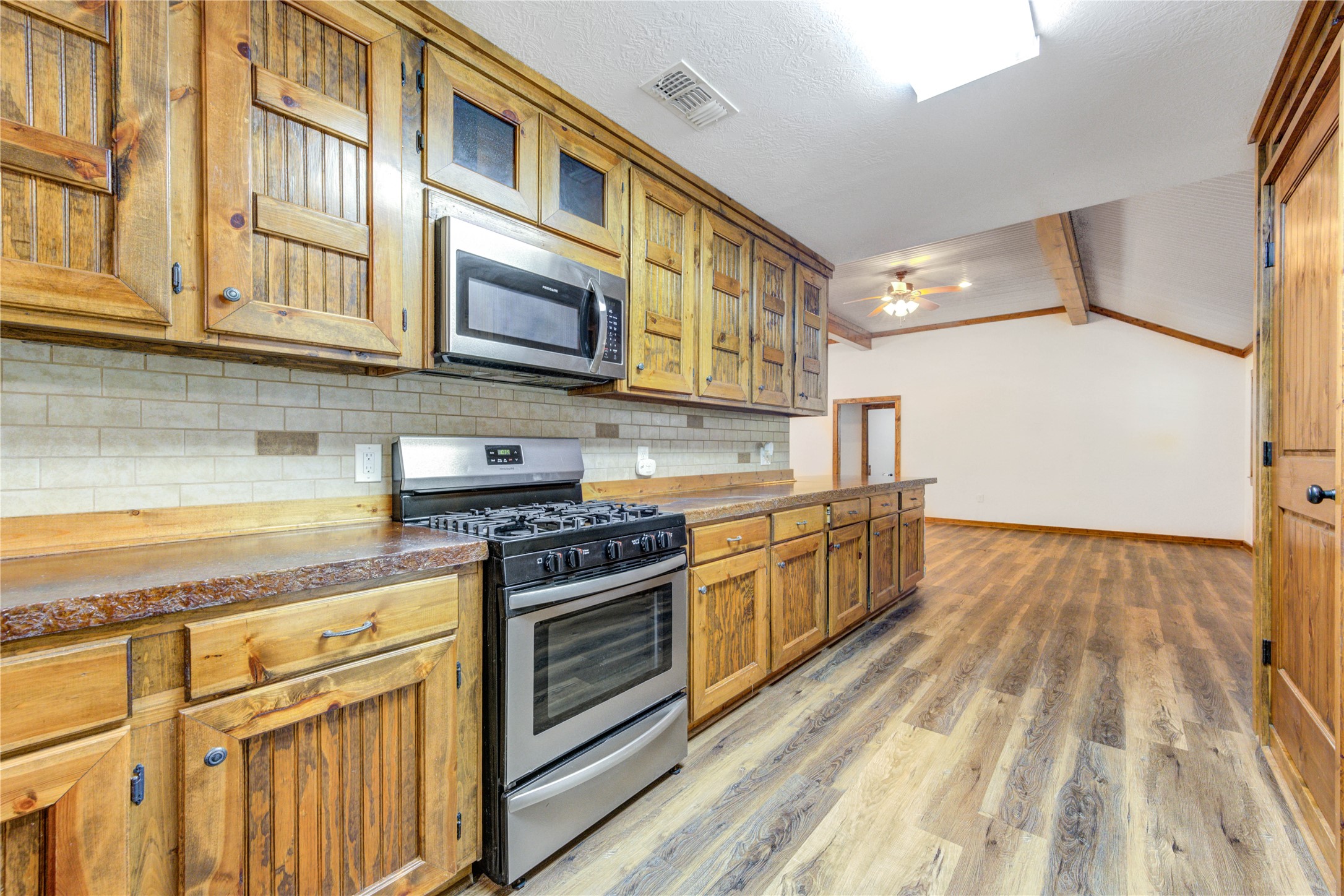 8805 Sun Oil Road Brenham, TX 77833 - Photo 22 of 50 Spacious kitchen with rustic wood cabinetry, modern stainless steel appliances, and a long countertop, leading into an open-concept living area with vaulted ceilings and wood flooring.
