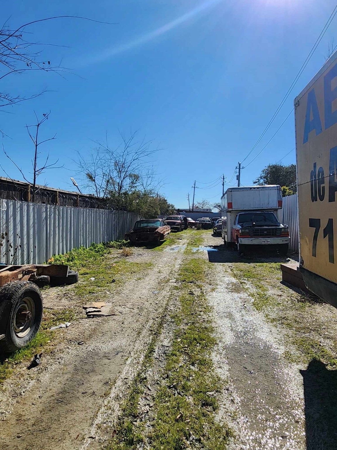 0 Rasmus Drive Houston, TX 77063 - Photo 11 of 11 This photo shows a narrow, unpaved driveway lined with metal fencing and vehicles, suggesting a storage or industrial area. The ground is grassy with some muddy patches, and there are a few scattered pieces of debris.