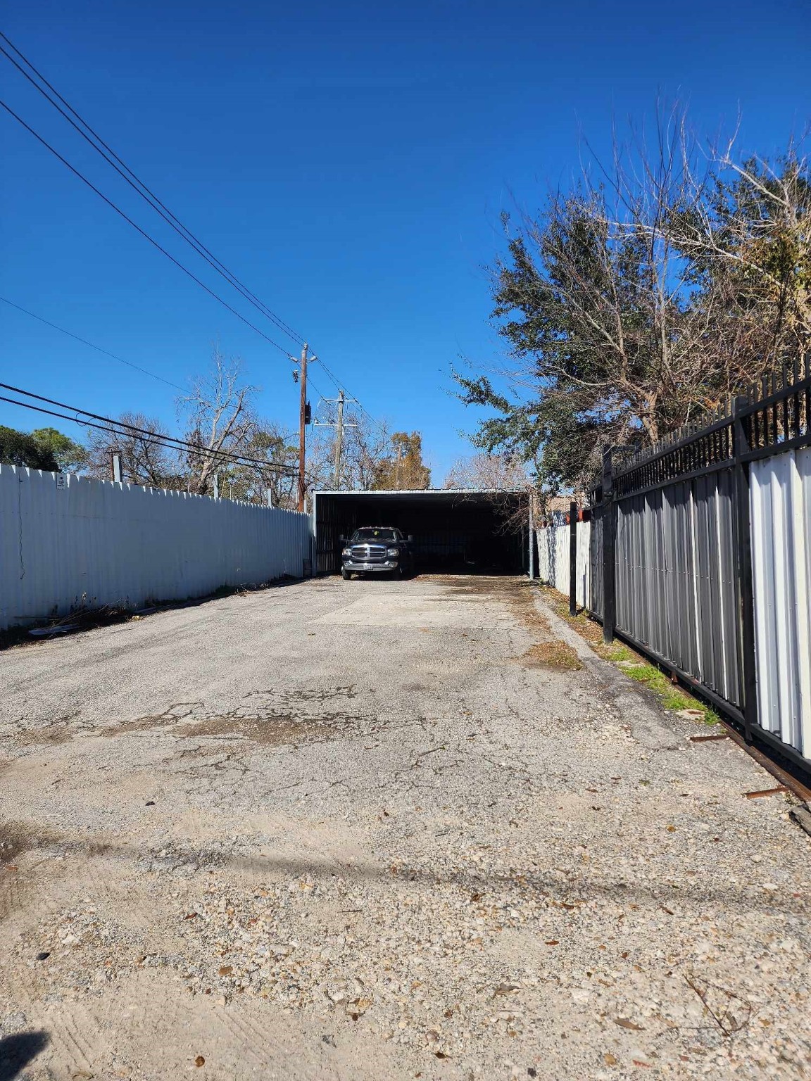 0 Rasmus Drive Houston, TX 77063 - Photo 4 of 11 This image shows a long driveway with a carport at the end, providing covered parking for vehicles. The area is enclosed by fences on both sides, featuring a mix of paved and gravel surfaces. The setting is open to the sky with some trees visible.