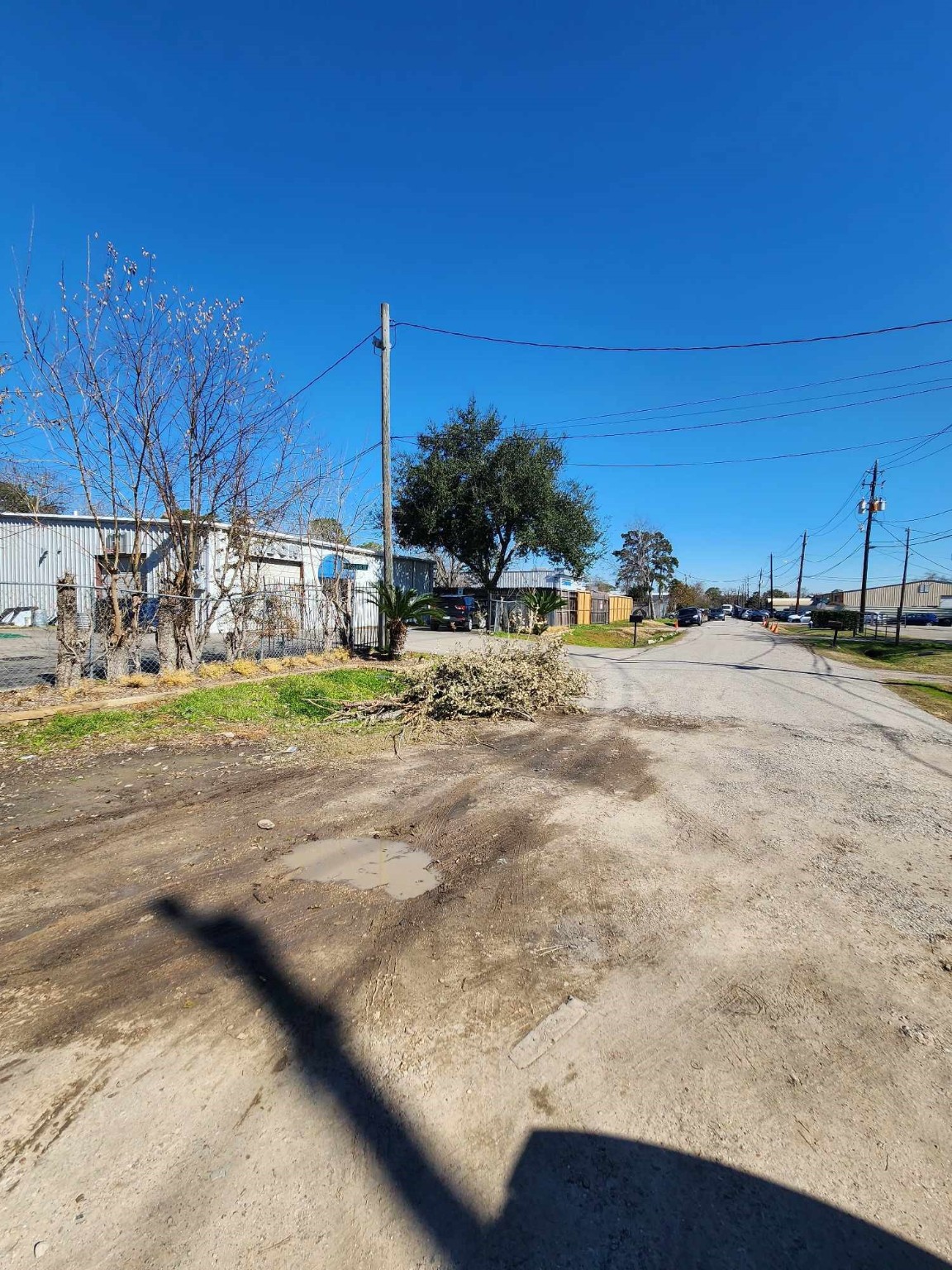 0 Rasmus Drive Houston, TX 77063 - Photo 5 of 11 This photo shows a quiet residential street with a mix of paved and unpaved surfaces. There are utility poles and wires lining the street, alongside some trees and vegetation. The area appears to be calm and open, offering a rural or semi-industrial feel.