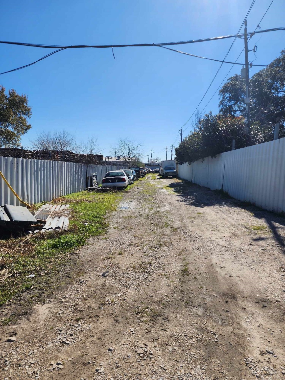 0 Rasmus Drive Houston, TX 77063 - Photo 6 of 11 The photo shows a narrow, unpaved alley lined with cars and bordered by tall, corrugated metal fences. The area appears somewhat cluttered, with some patches of grass and debris along the sides. It’s a sunny day with clear skies.