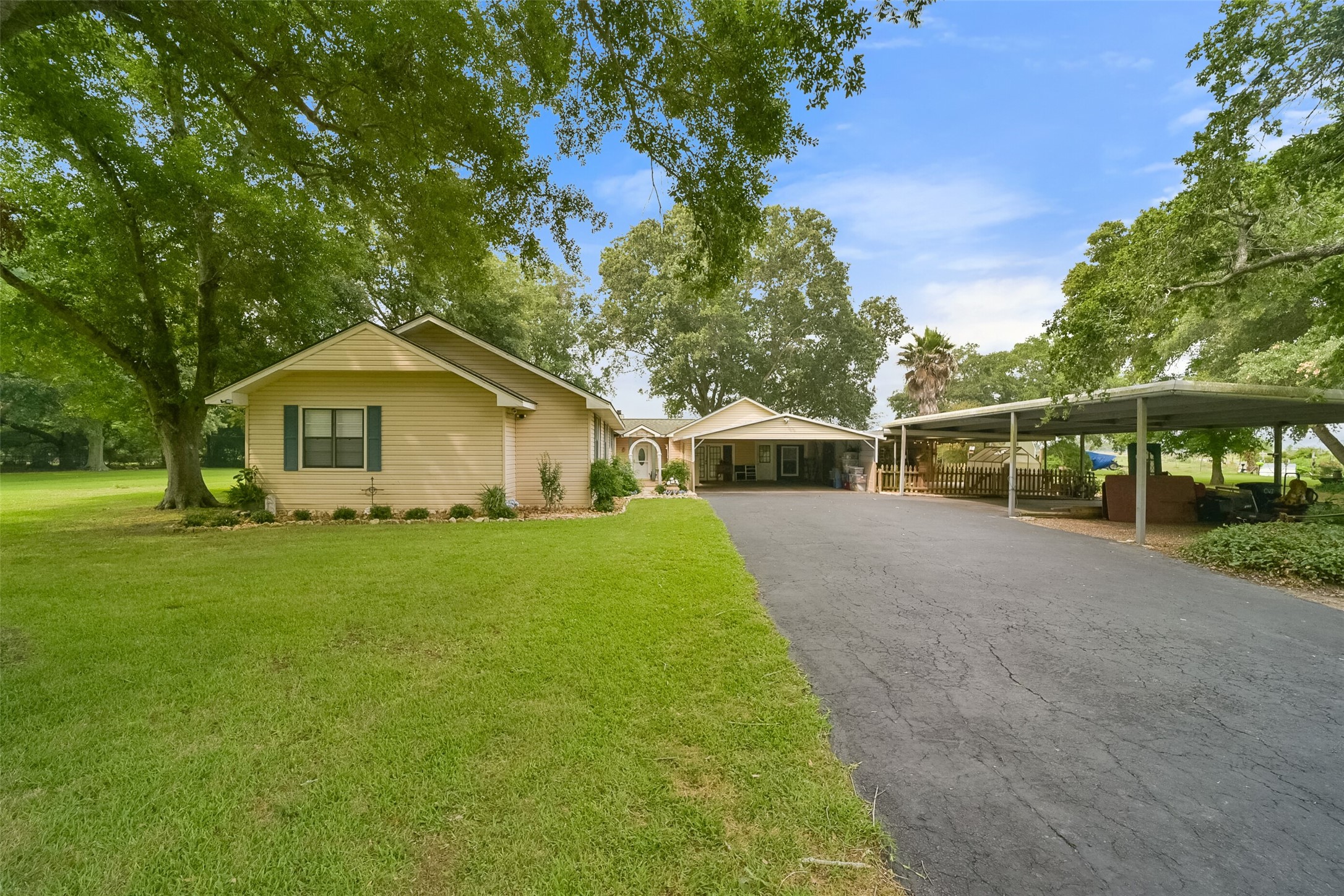 This single-story home features a large front yard with mature trees, a paved driveway leading to a carport, and a cozy porch, all set in a spacious and green setting.