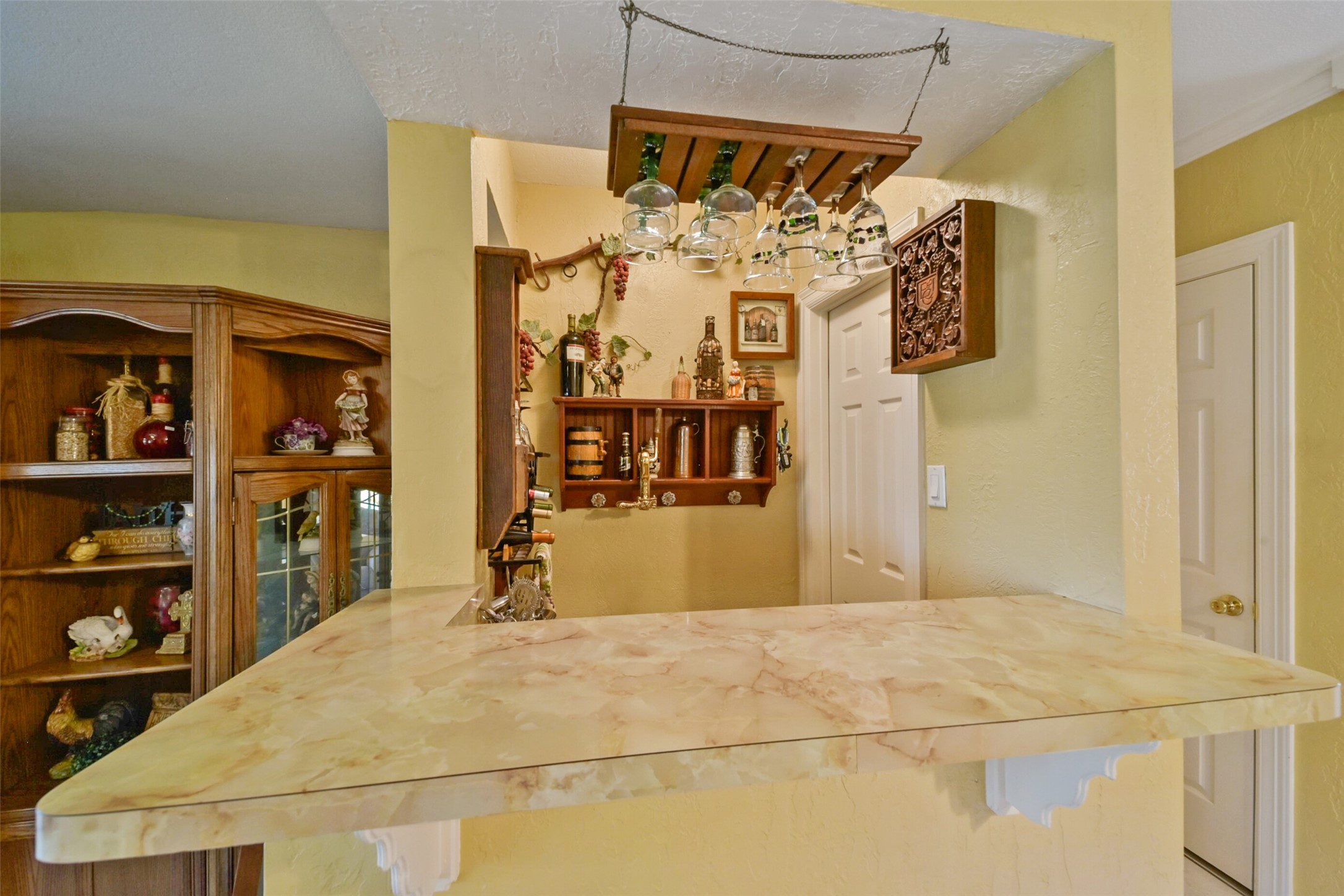 13710 Mueck Road Needville, TX 77461 - Photo 14 of 42 Experience the charm of this cozy kitchen nook with a sleek marble countertop, elegant wooden cabinetry, and a stylish hanging wine glass rack. Bright and tastefully decorated, it offers a warm and inviting atmosphere.