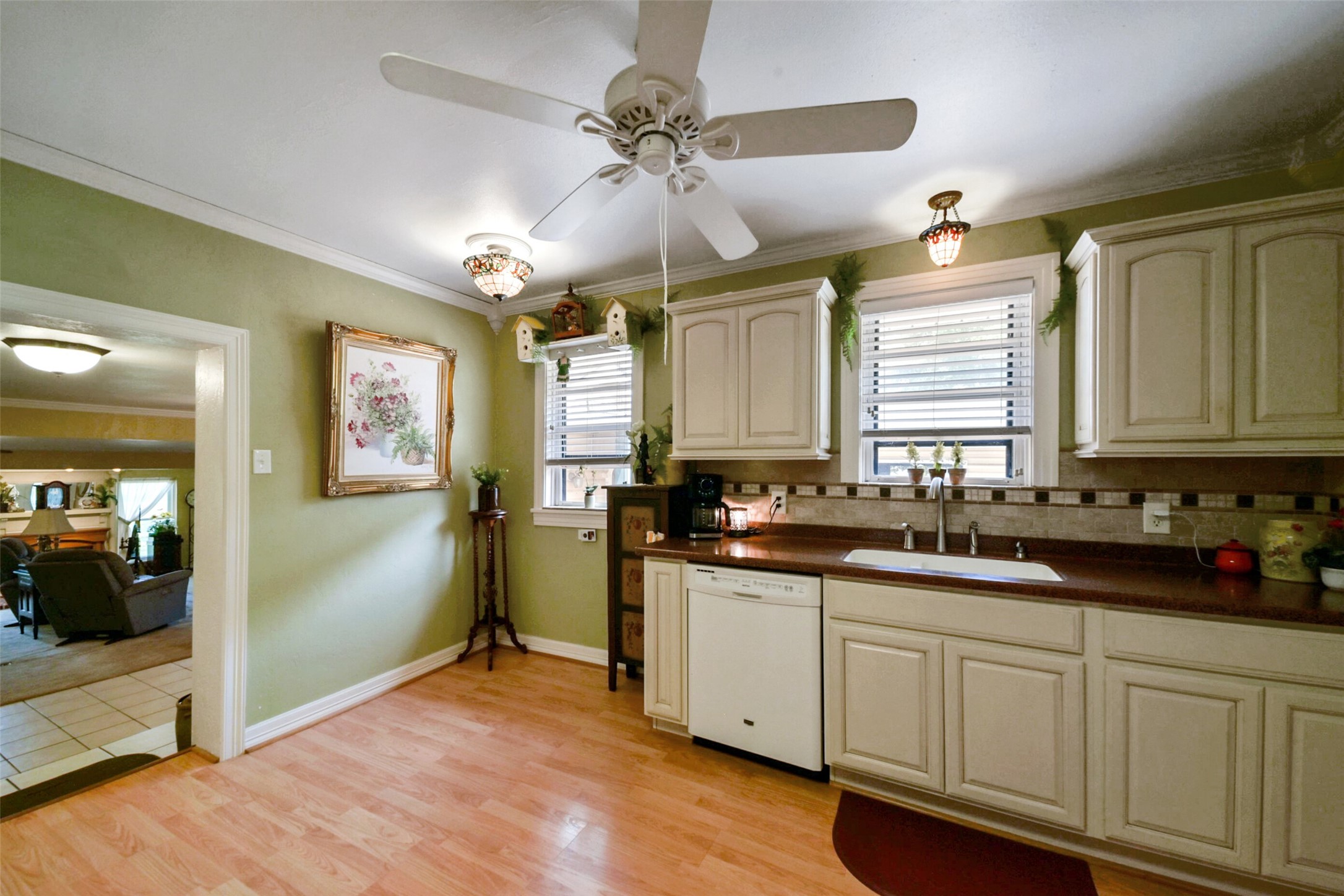 13710 Mueck Road Needville, TX 77461 - Photo 19 of 42 This is a cozy kitchen featuring cream-colored cabinetry, laminate countertops, and a tiled backsplash. It includes a double sink with a window above it, white appliances, and hardwood flooring. The room is lit with a ceiling fan and two pendant lights, and there's an open doorway leading to the adjacent living area.