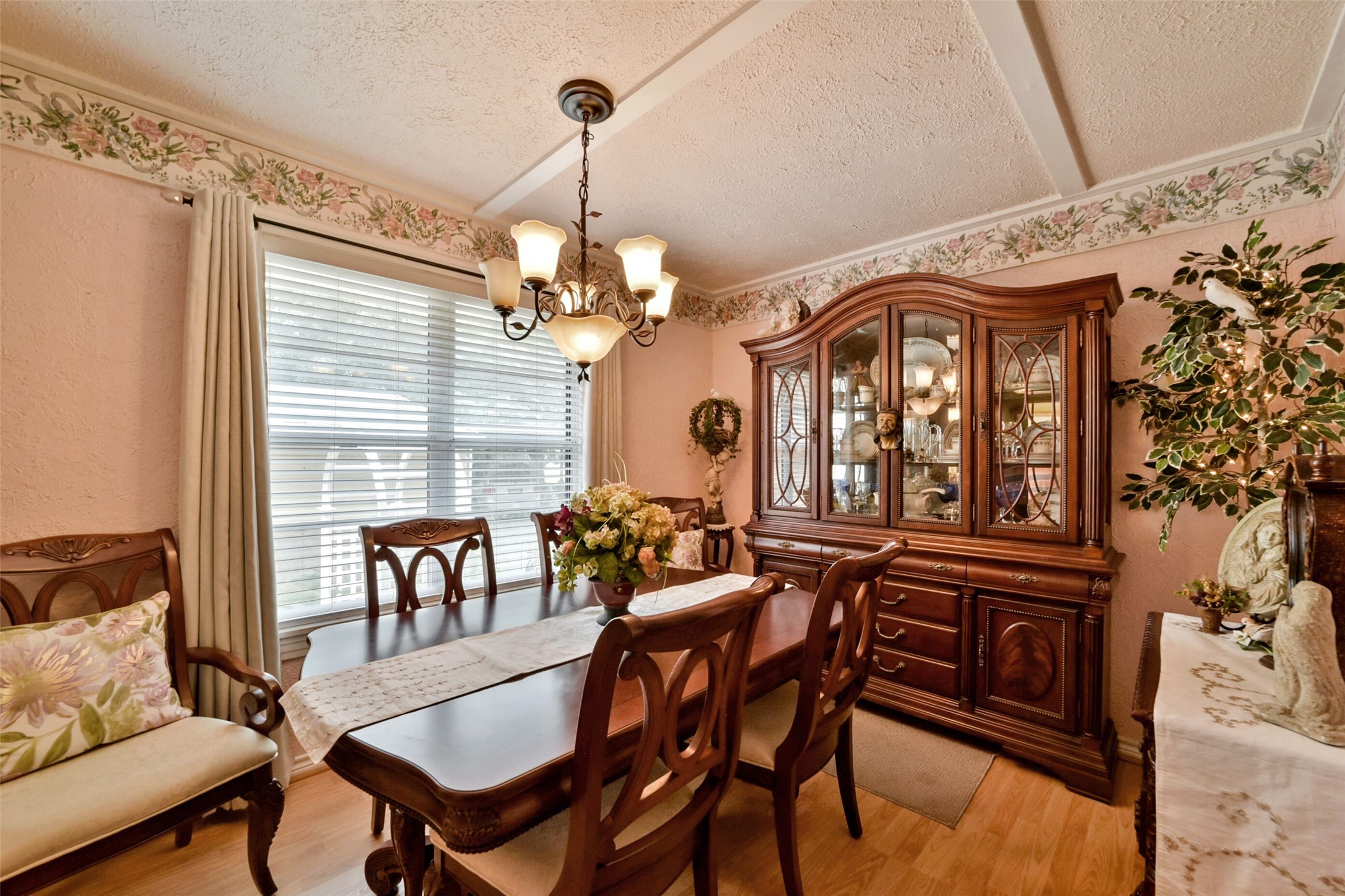 13710 Mueck Road Needville, TX 77461 - Photo 20 of 42 This is a cozy, traditional dining room featuring an elegant wooden table with matching chairs, a classic china cabinet, and a chandelier. Natural light filters in through the blinds of a large window, and the room is finished with decorative floral wallpaper borders.
