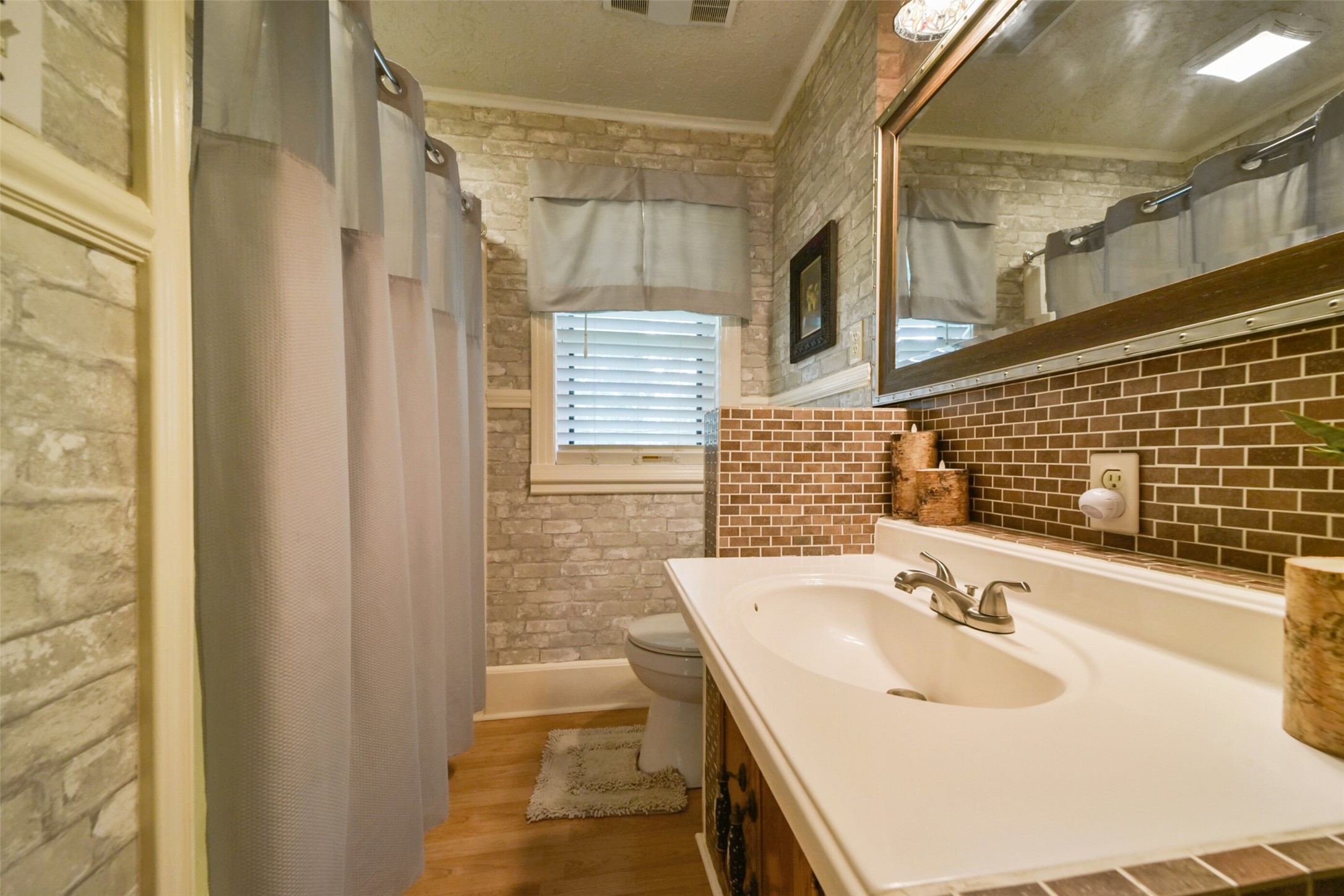 13710 Mueck Road Needville, TX 77461 - Photo 24 of 42 This is a cozy bathroom featuring a stone accent wall, a brown subway tile backsplash, and a single white vanity with undermount sink. There's a shower with a semi-transparent curtain and a toilet area, complemented by neutral tones and a window providing natural light.