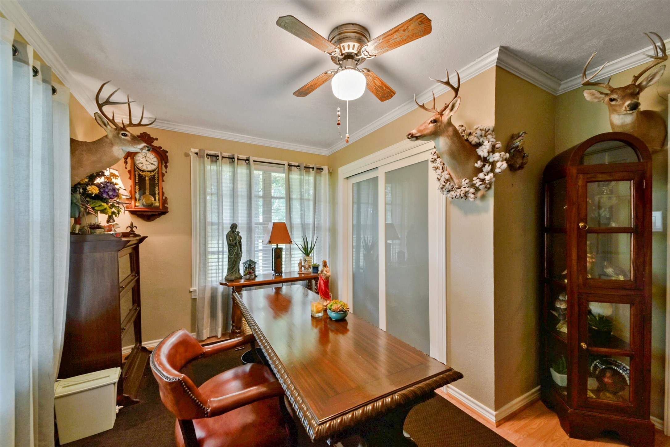 13710 Mueck Road Needville, TX 77461 - Photo 25 of 42 This room features a traditional decor with mounted deer heads on the walls, a wooden desk and chair set, a ceiling fan, and a glass-fronted display cabinet. The space is well-lit, with natural light coming through the windows dressed in neutral curtains.