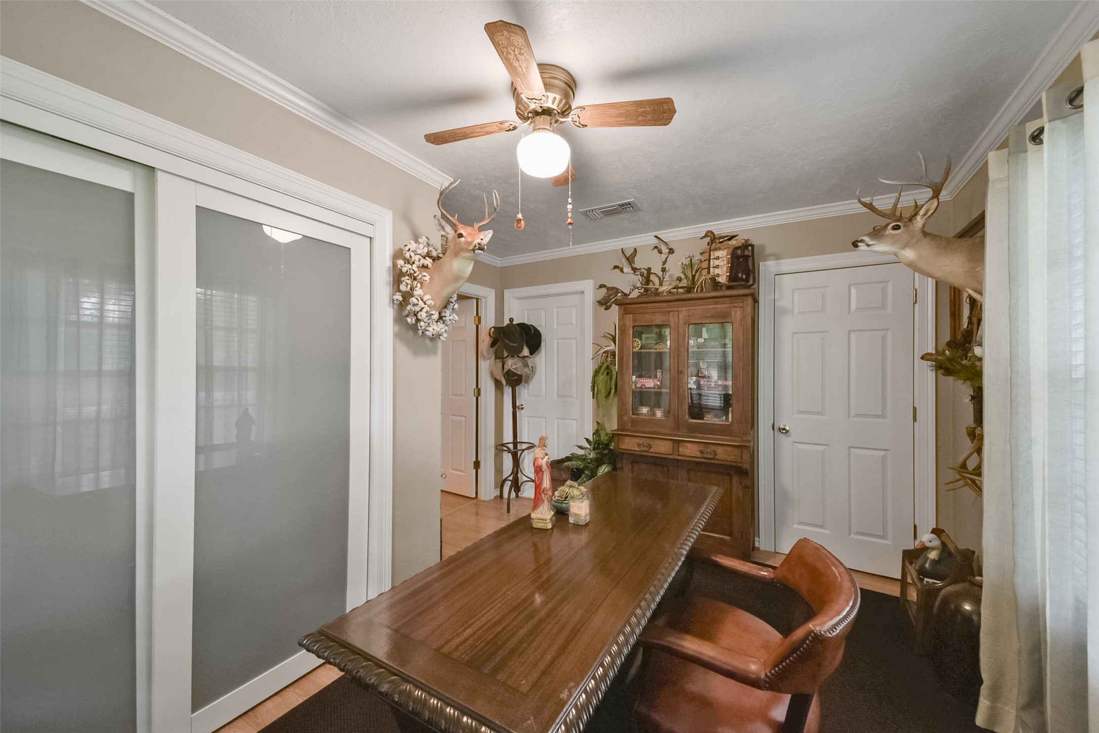 13710 Mueck Road Needville, TX 77461 - Photo 26 of 42 This room features neutral walls, wood furniture, and a ceiling fan. The space includes a dining table with leather chairs and is adorned with wildlife-themed decor, including mounted deer heads. It has glass-paneled doors and a window with sheer curtains, contributing to the room's natural light.