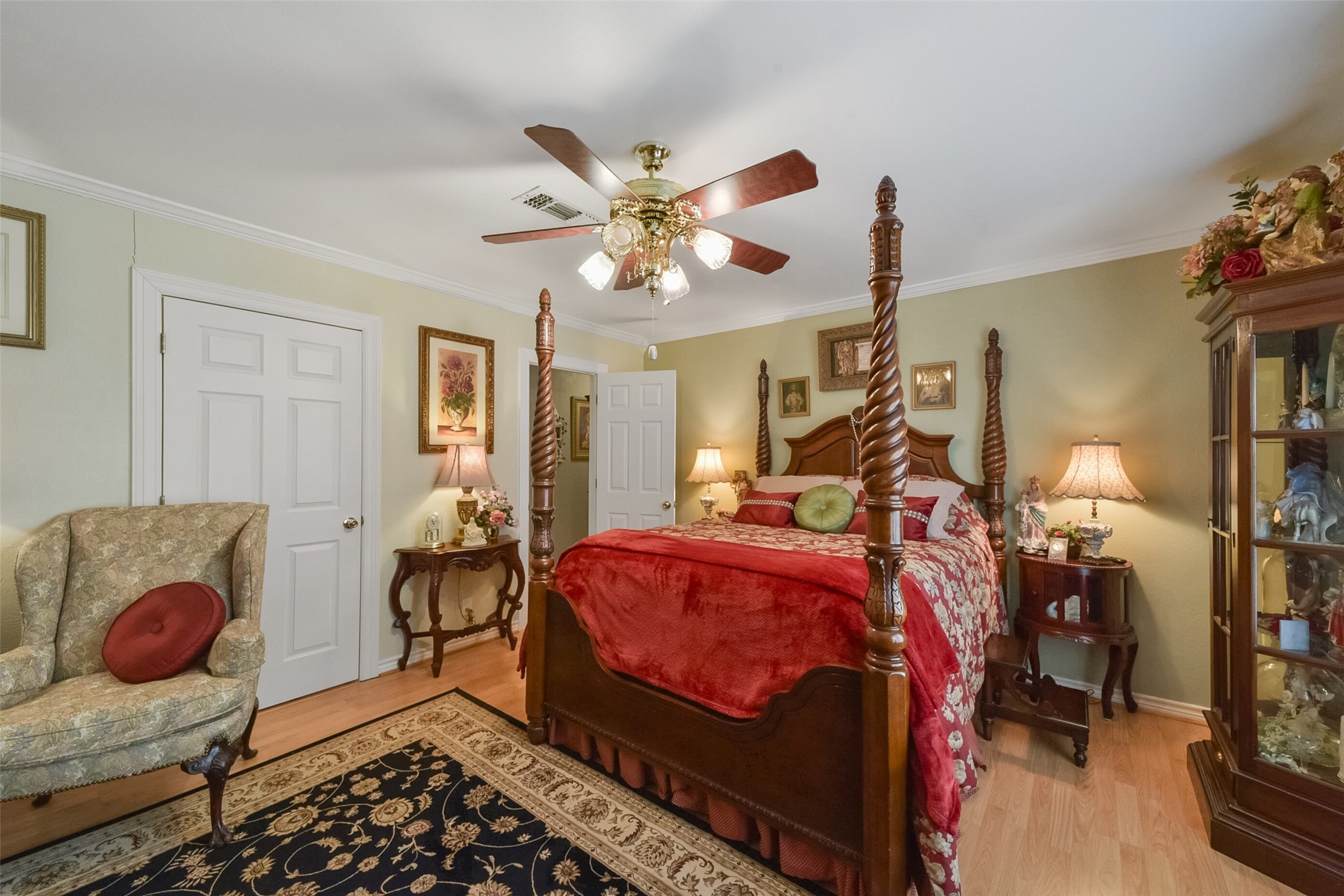 13710 Mueck Road Needville, TX 77461 - Photo 28 of 42 This is a traditional-style bedroom featuring a four-poster bed with a red bedspread, hardwood floors, a ceiling fan, and classic decor. The room includes a comfortable armchair, side tables with lamps, and a display cabinet. It's well-maintained with a cozy and elegant atmosphere.