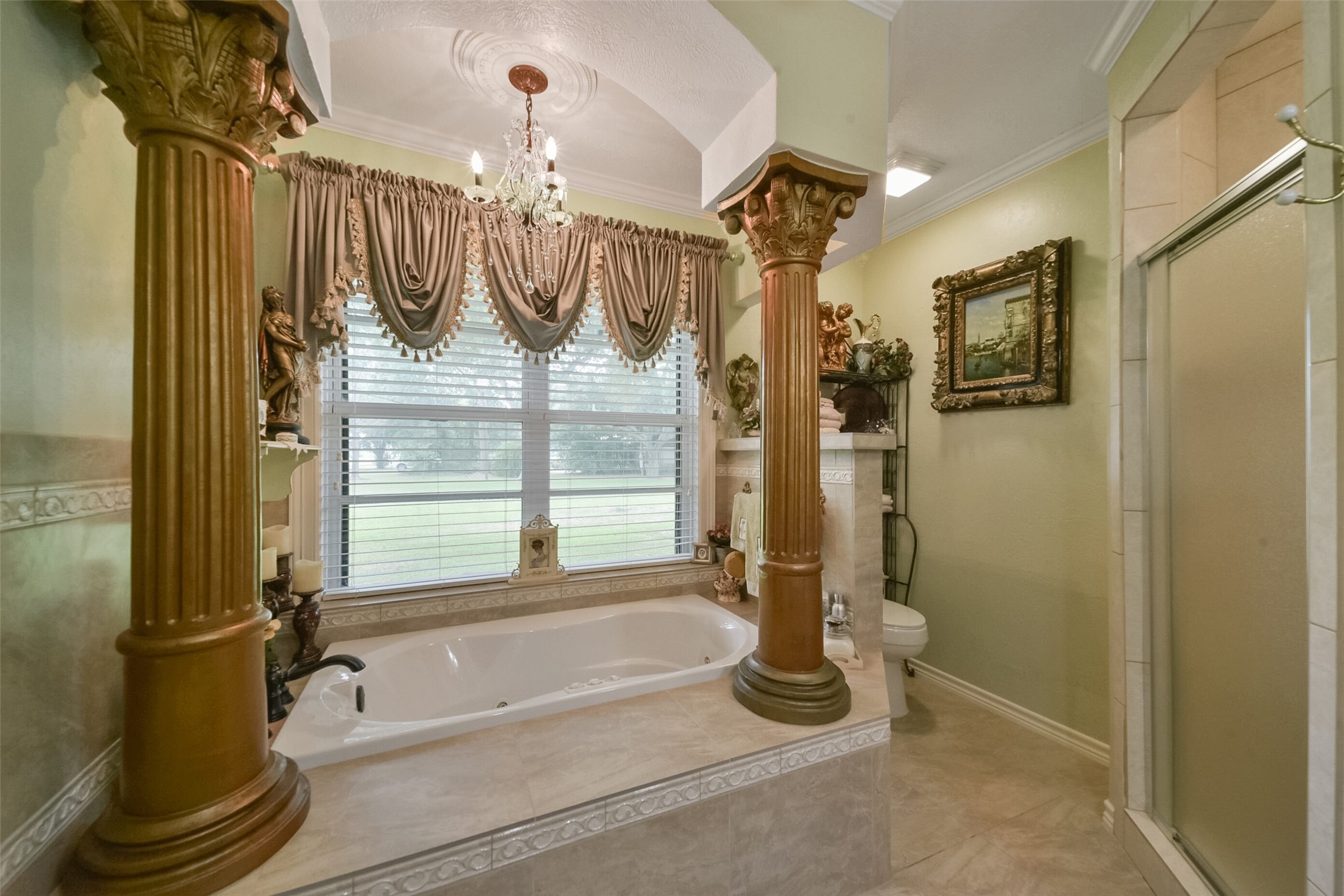 13710 Mueck Road Needville, TX 77461 - Photo 31 of 42 This is an ornate bathroom featuring a large bathtub with luxurious columns on either side, elegant drapery, a chandelier, and a separate shower stall. Natural light comes in through a window overlooking the yard.