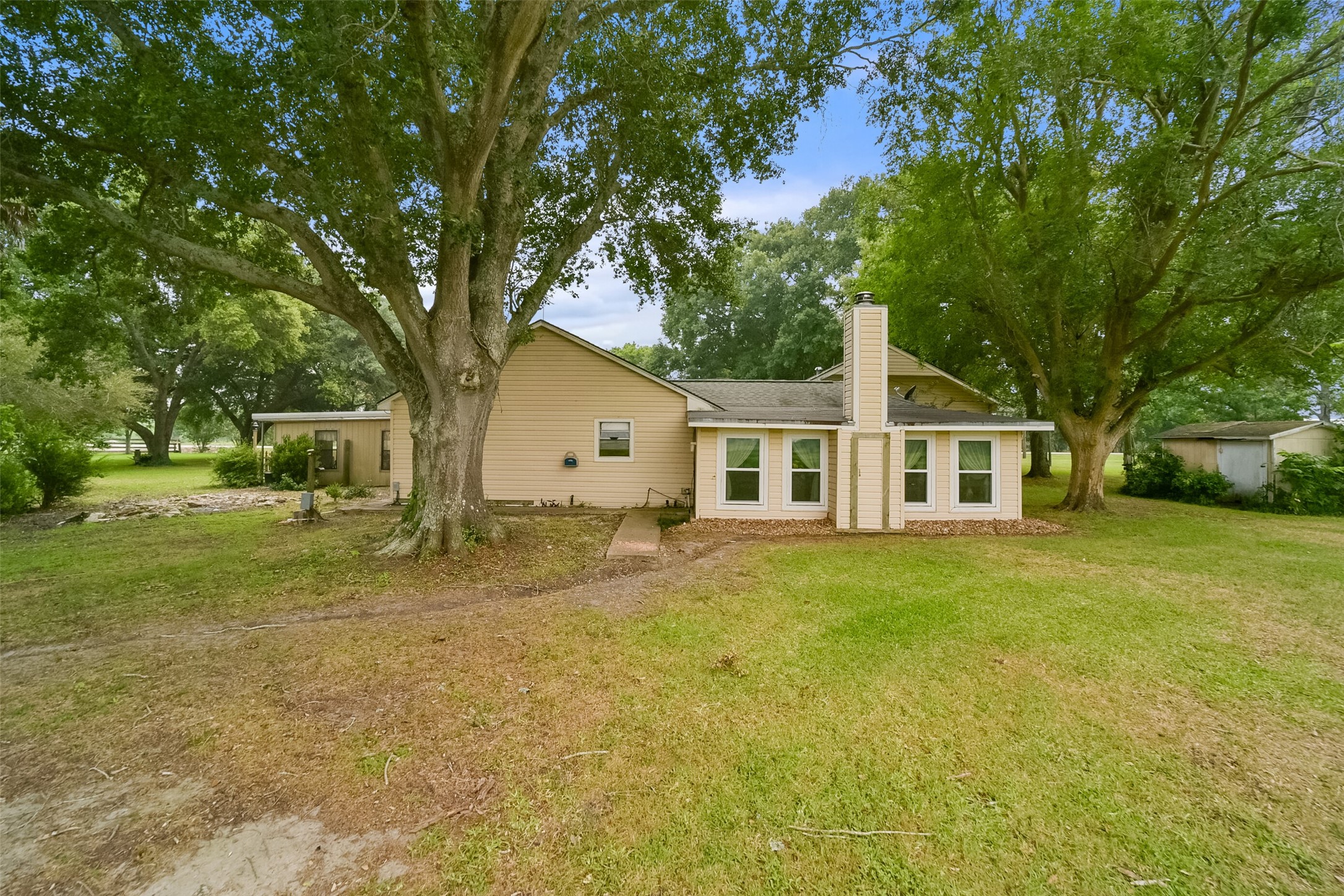 13710 Mueck Road Needville, TX 77461 - Photo 34 of 42 This is a single-story home featuring a beige exterior with white trim, a prominent chimney, and large windows. The property boasts mature trees and a spacious yard.