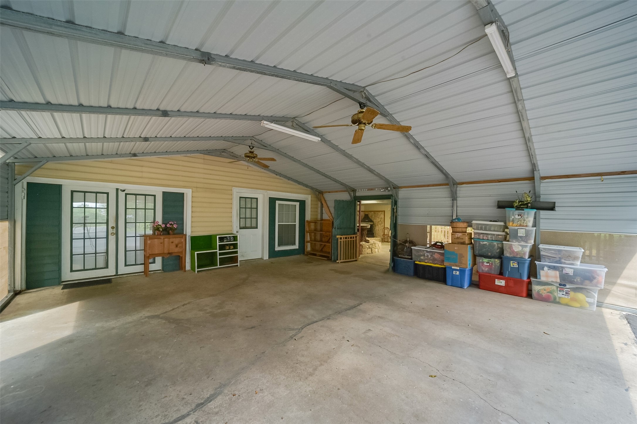 13710 Mueck Road Needville, TX 77461 - Photo 40 of 42 This is a spacious, semi-enclosed garage area with a high metal ceiling and two ceiling fans. It features concrete flooring, storage space along one wall, and dual French doors leading into the home. The space is utilitarian, with ample room for parking, storage, or potentially a workshop area.