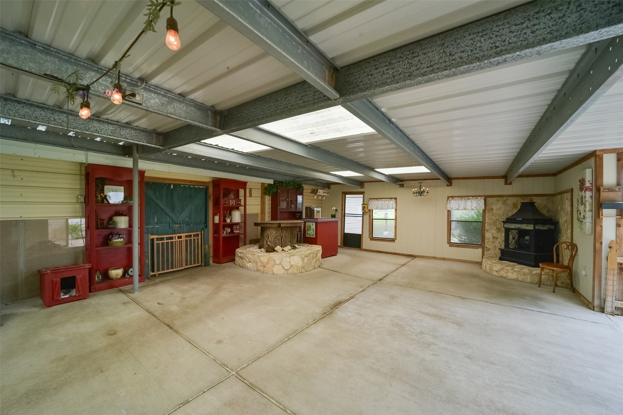 13710 Mueck Road Needville, TX 77461 - Photo 41 of 42 This is a spacious, partially enclosed garage or carport area featuring a cozy corner with a stone fireplace, a built-in bar, and decorative shelving. The space is versatile, with ample room for parking or entertaining, and it has a rustic charm enhanced by string lights overhead.