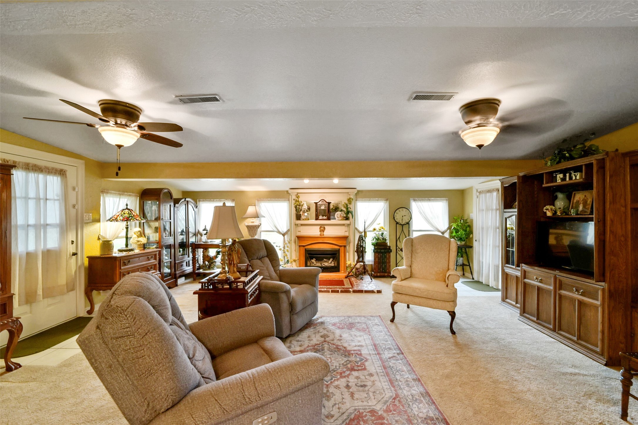 13710 Mueck Road Needville, TX 77461 - Photo 10 of 42 Experience the warmth of this spacious living room, featuring a cozy fireplace, comfortable seating, ceiling fans, and a built-in bookcase. Its carpeted floors and abundant natural light create an inviting atmosphere.